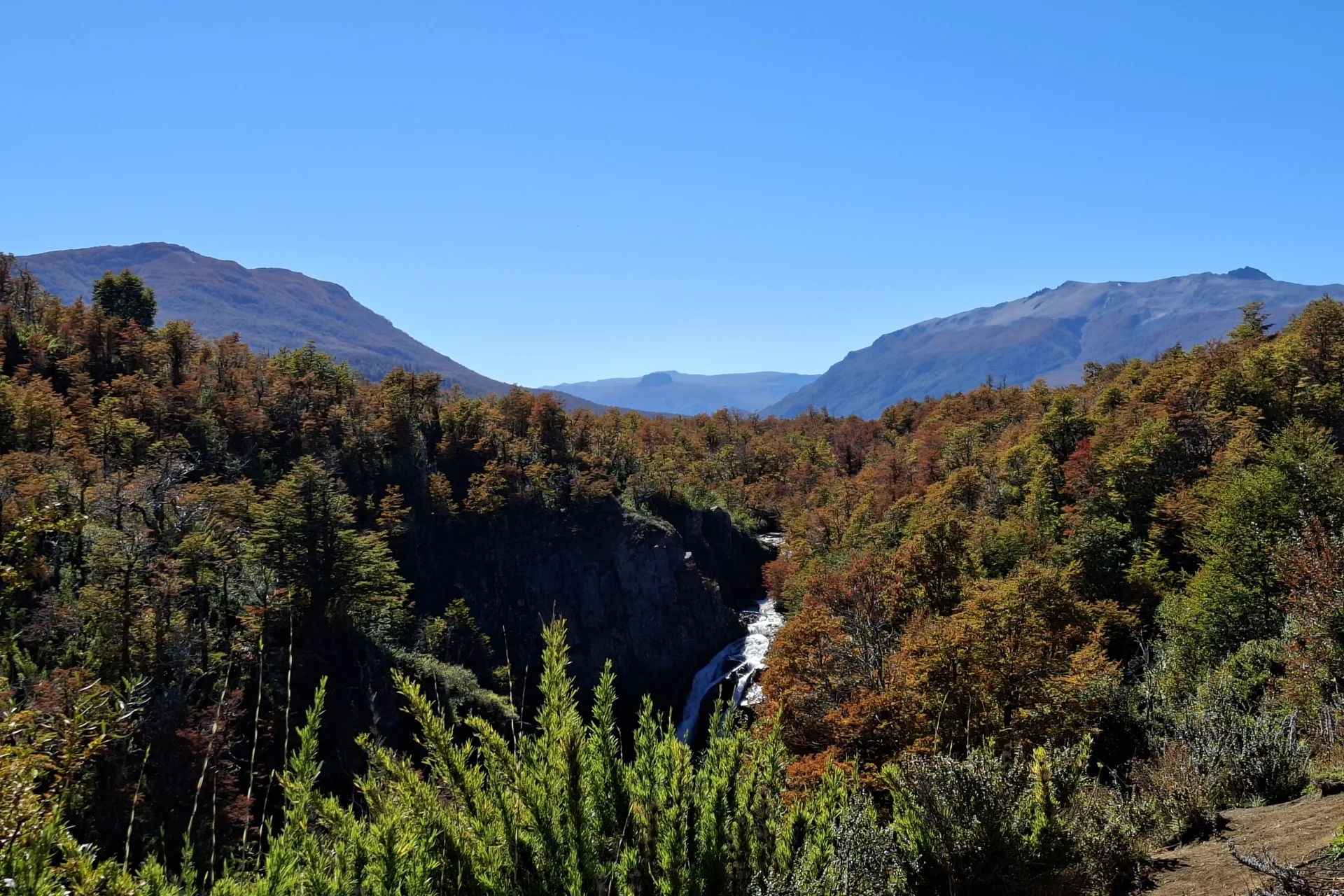 En la imatge situada horitzontalment, s’hi observa unes muntanyes al darrere, amb uns arbres de colors tardor i una cascada en primer terme.