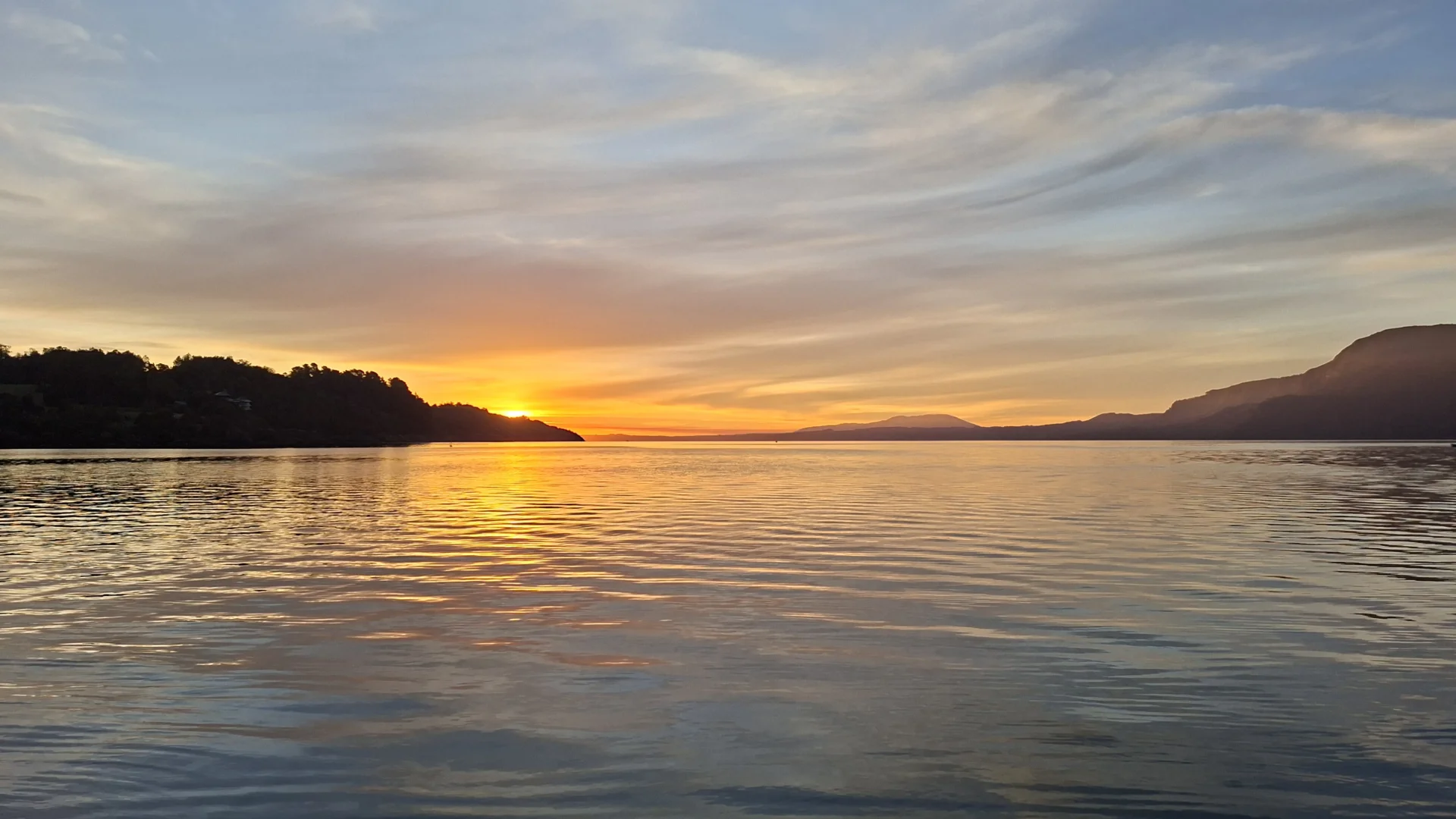 Posta de sol sobre el llac Villarrica a Pucón, Xile. El sol es pon darrere d'un turó a l'esquerra, generant reflexos de color taronja i groc sobre l'aigua. A l'extrema dreta es perfila la silueta del volcà Villarrica sota un cel amb núvols lineals de tons lila i blau.