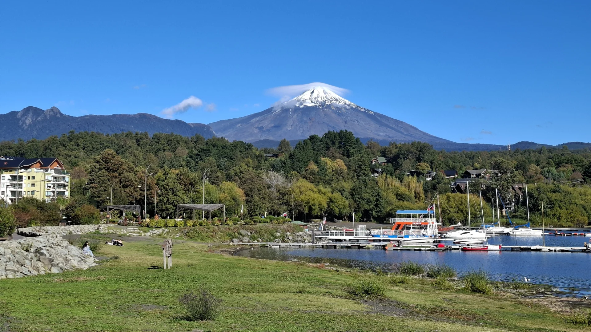 Vista del volcà Villarrica amb el cim nevat i un núvol lenticular superior sota un cel blau clar. En primer pla, una zona de gespa i un espigó de pedres a la riba del llac. A la dreta, un moll amb embarcacions amarrades, incloent-hi un catamarà de colors vermell i blau.