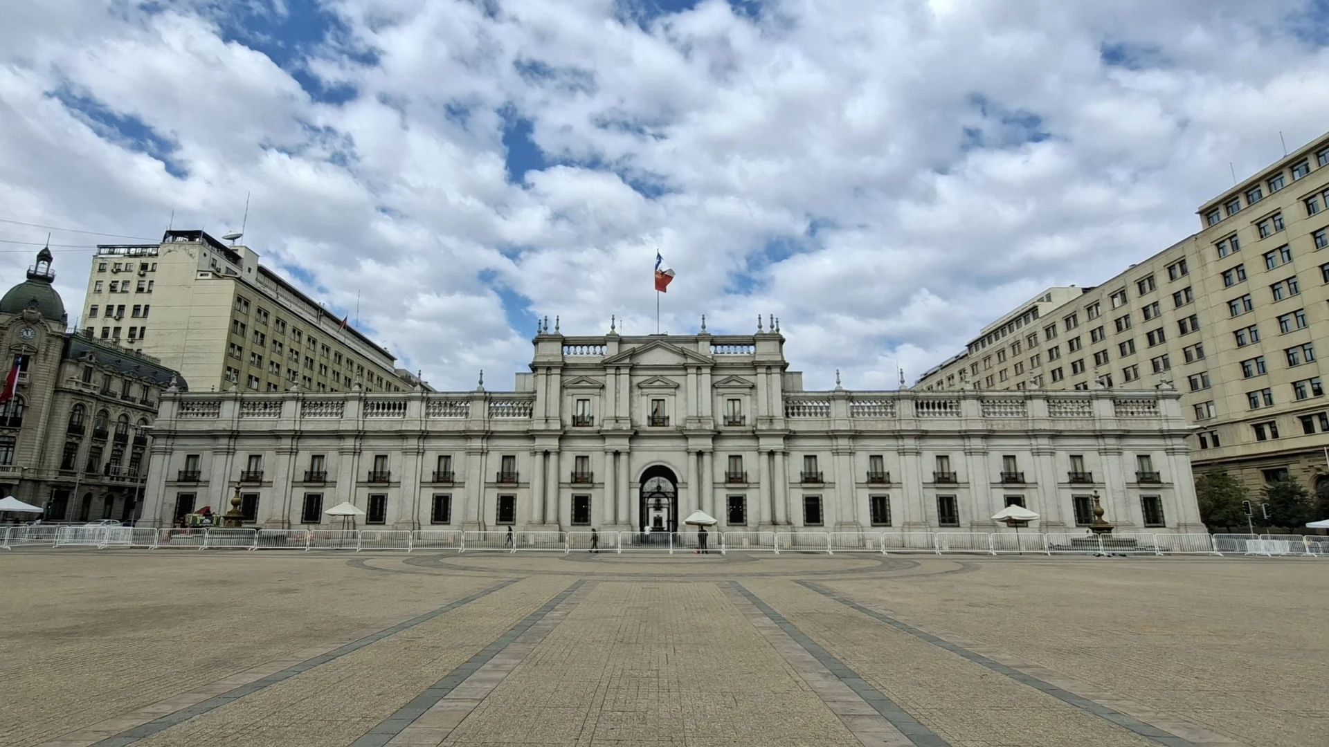 Vista de la façana del Palau de La Moneda a Santiago de Xile, sota un cel parcialment ennuvolat. L'edifici neoclàssic central de color clar té una bandera a la part superior central. Al davant, hi ha una gran plaça pavimentada i buida, delimitada per tanques de seguretat temporals i amb para-sols moderns de color clar. Edificis urbans més alts envolten la plaça.