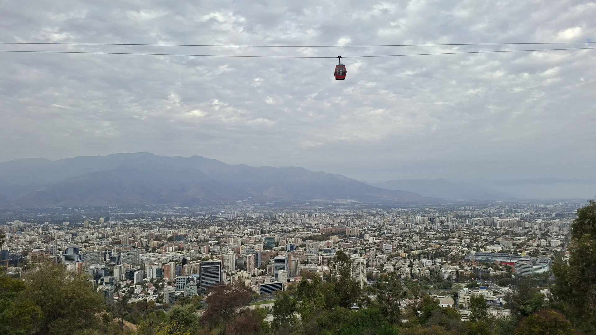Vista panoràmica de la ciutat de Santiago de Xile des del turó de San Cristóbal. Una cabina de telefèric de color vermell penja dels cables a la part superior central. Al fons, es veu la silueta de la serralada dels Andes sota un cel gris i ennuvolat amb boirina. En primer pla, apareix la vegetació del turó que dona pas a una densa trama d'edificis urbans.