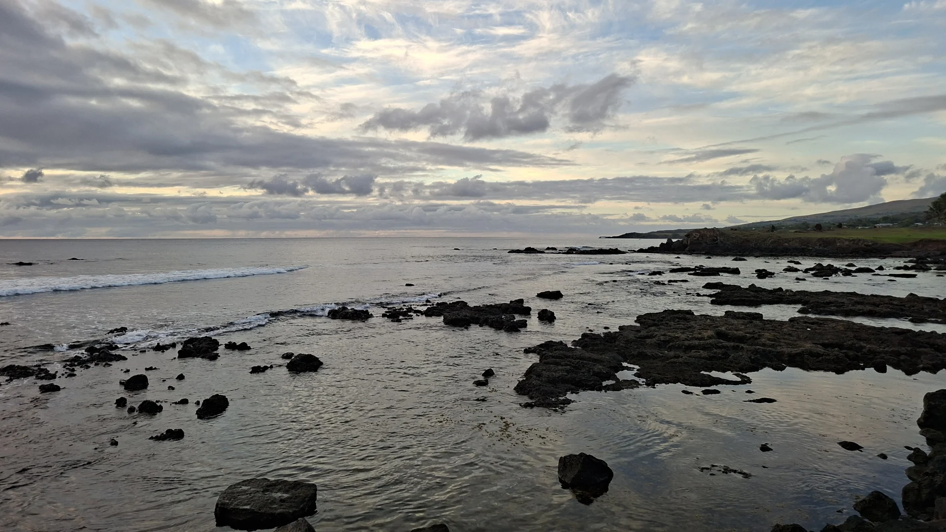 Paisatge costaner a Rapa Nui durant el capvespre sota un cel molt ennuvolat. En primer pla, l'aigua del mar amb roques volcàniques negres escampades i una petita onada a l'esquerra. A l'horitzó s'aprecien clarianes de tons grocs i taronges que es reflecteixen sobre l'aigua.