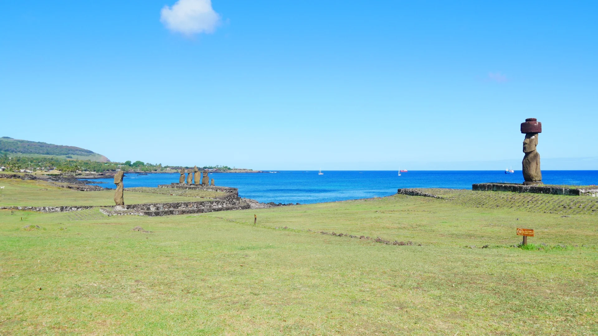 Vista panoràmica del complex arqueològic de Tahai a Rapa Nui, amb el Moai solitari d'Ahu Ko Te Riku i el seu tocat vermell (pukao) a la dreta, i una altra plataforma amb cinc Moais al fons a l'esquerra, davant d'un mar blau intens sota un cel clar.