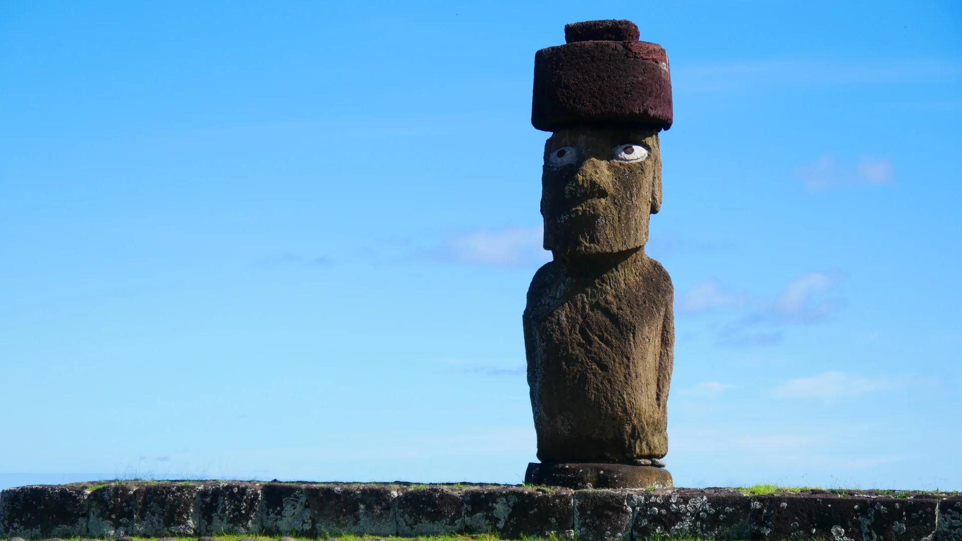 Fotografia d'un moai a Rapa Nui, a la zona de Tahai, amb ulls de corall i un pukao vermell al cap. L'estàtua de pedra mira endavant sota un cel blau clar, dret sobre una plataforma de pedra a la base.