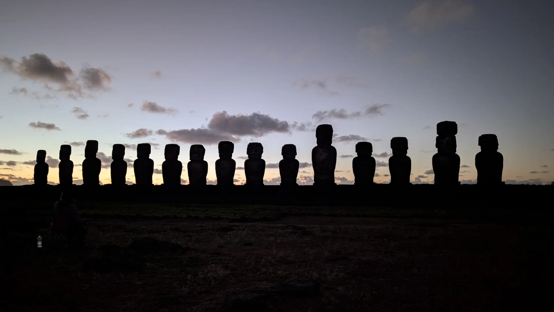 Silueta de la plataforma d'Ahu Tongariki amb els quinze Moais alineats, retallada contra el cel durant la sortida del sol, que mostra una franja de tons taronja i groc a l'horitzó que es difumina cap al blau.