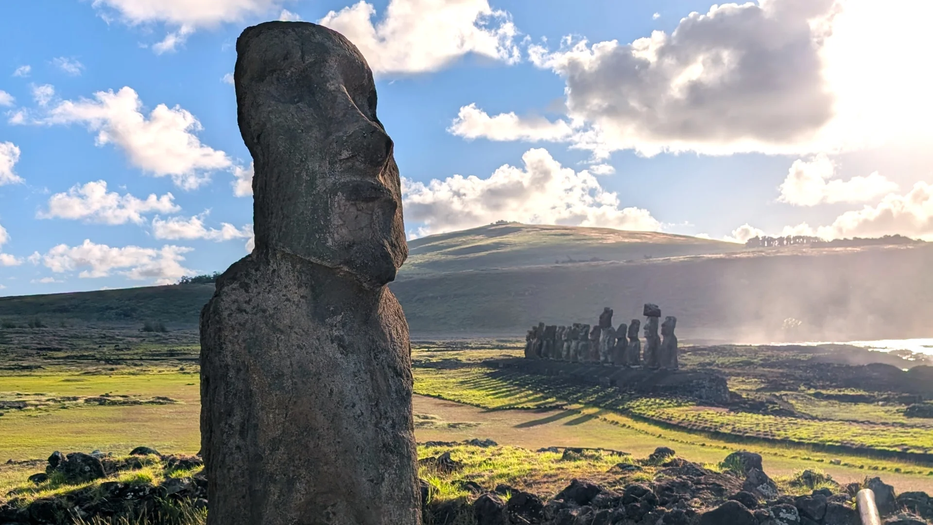 Primer pla de perfil d'un Moai de pedra a l'esquerra de la imatge. Al fons, enmig d'un gran prat verd il·luminat per la llum del matí, es distingeix la plataforma d'Ahu Tongariki amb els quinze Moais alineats que projecten llargues ombres, sota un cel blau amb núvols blancs i un turó al fons.