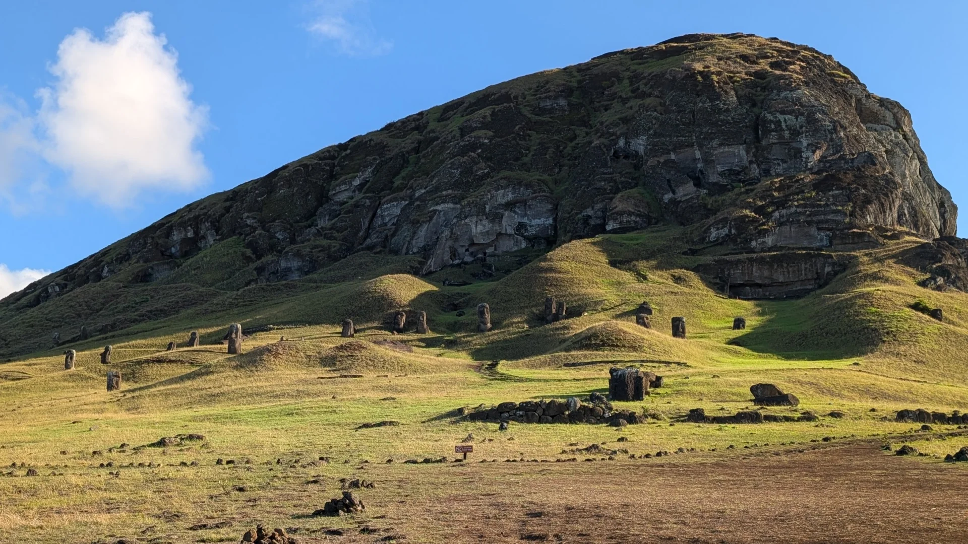 Vista de la gran muntanya de la pedrera de Rano Raraku a Rapa Nui, amb nombrosos Moais escampats i dempeus a mig enterrar pels turons de gespa verda il·luminats pel sol, sota un cel blau amb un gran núvol blanc a l'esquerra.