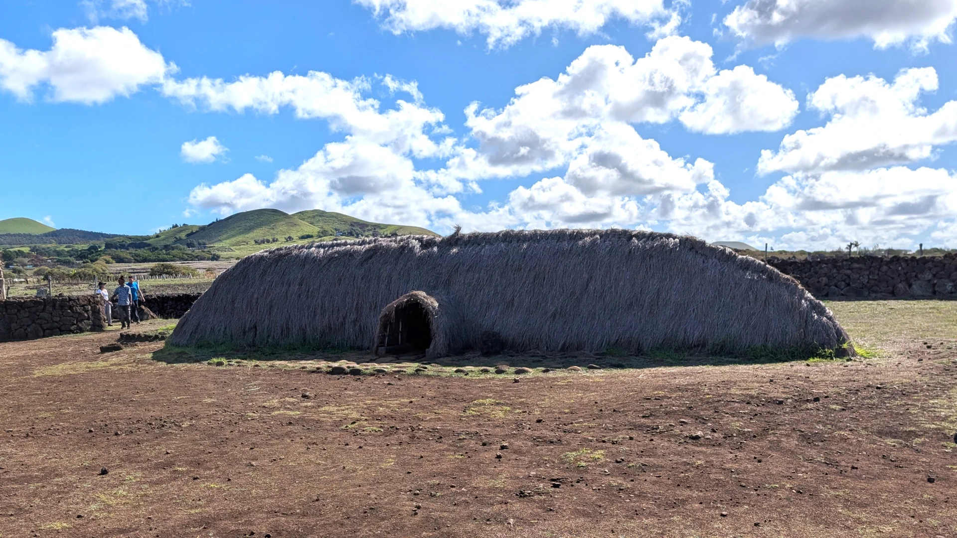 Una reproducció d'una Hare Vaka (casa-bot) tradicional de Rapa Nui a Akahanga, construïda amb palla i amb la seva característica forma de barca capgirada, mostrant una petita i baixa entrada arquejada al centre. Al fons es distingeixen turons verds sota un cel blau ple de núvols blancs.