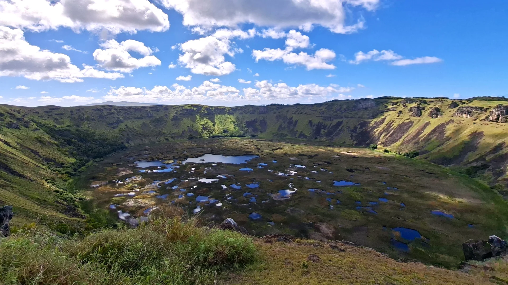 Vista panoràmica de l'immens cràter del volcà inactiu Rano Kau a Rapa Nui, amb parets escarpades de color verd que encerclen el fons, on es formen múltiples basses d'aigua blava entre zones de vegetació, sota un cel blau amb núvols blancs.