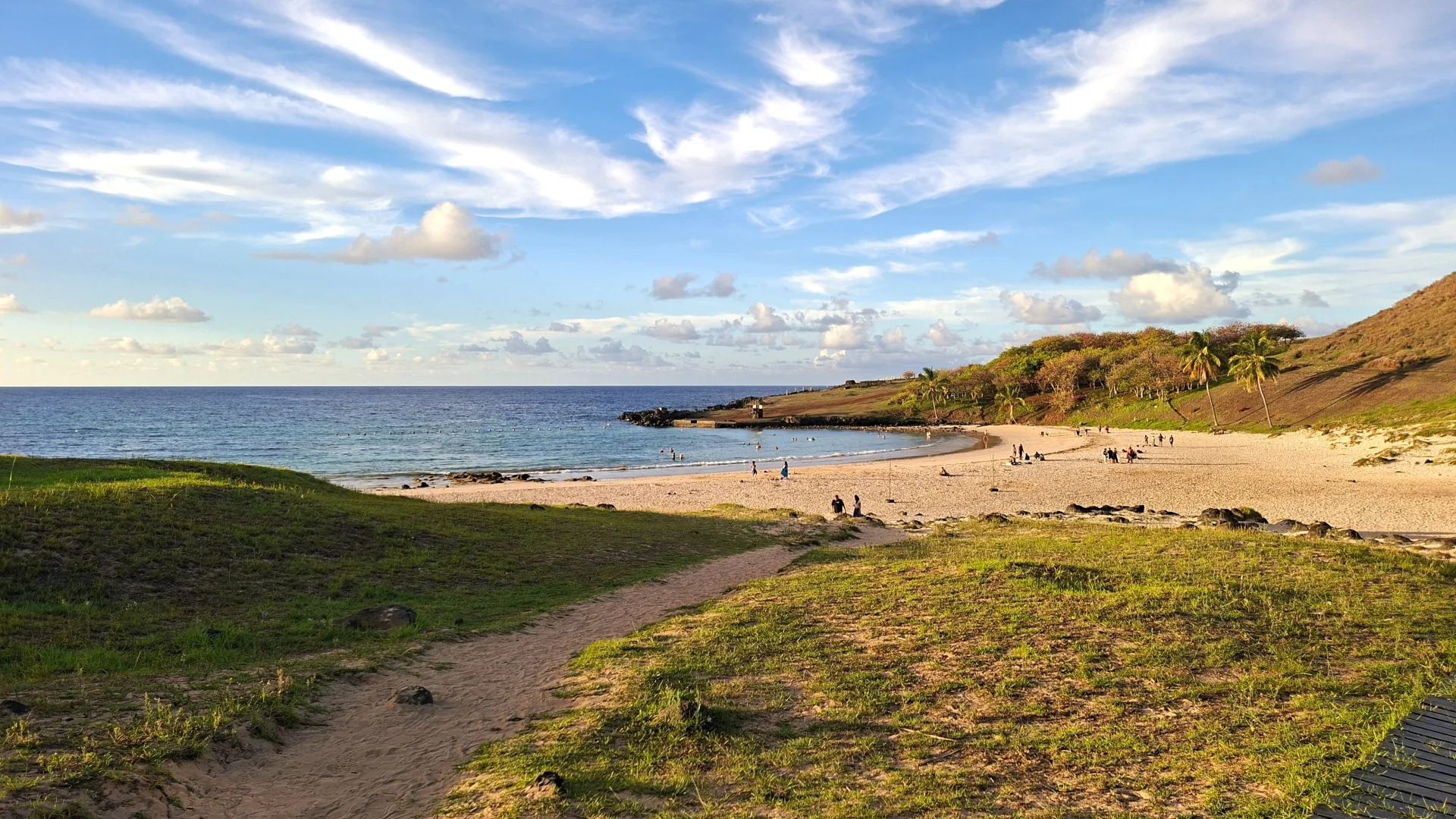 Vista de la platja d'Anakena a Rapa Nui des d'un camí de sorra que baixa entre turons de gespa verda. Es veu la badia de sorra clara amb petites siluetes de gent, un grup de palmeres a la banda dreta, l'oceà blau a l'horitzó i un cel ampli amb núvols fins i estriats il·luminats per la llum de la tarda.