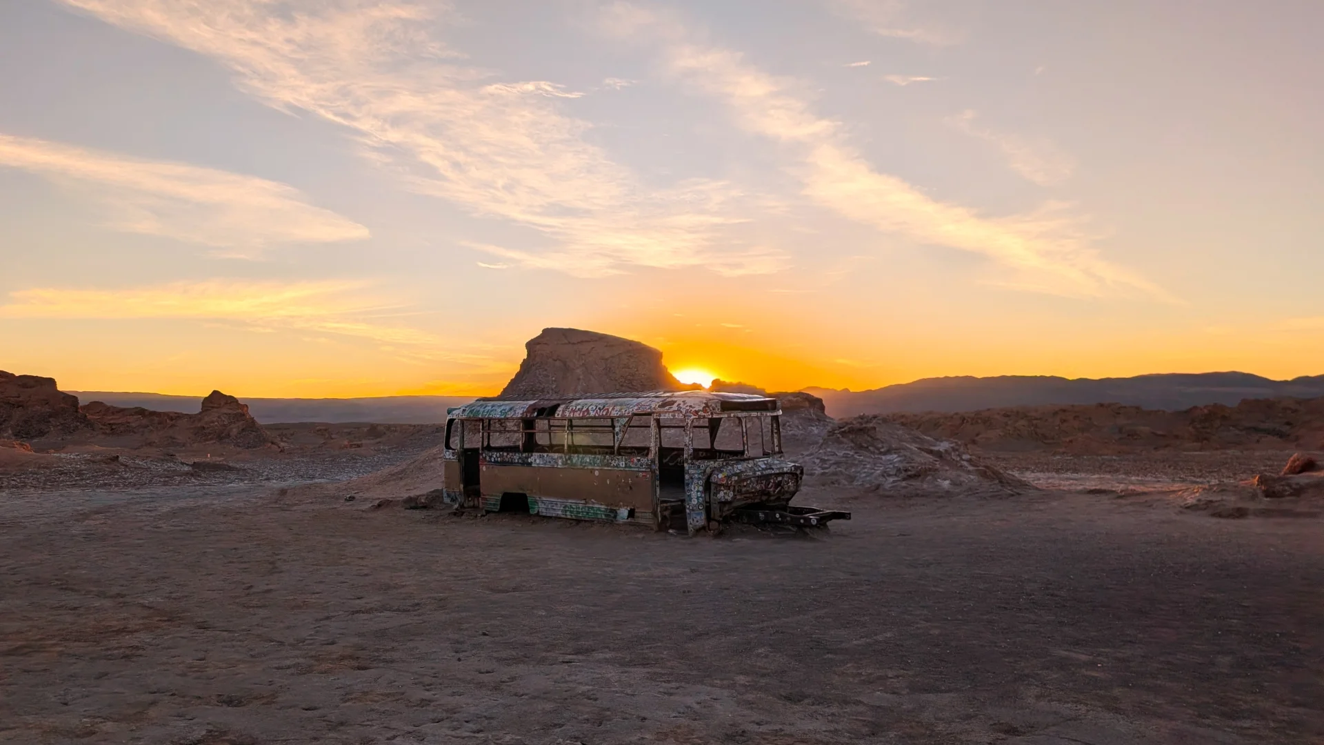 Un autobús vell i abandonat, cobert de grafits, enmig de l'àrid paisatge del desert d'Atacama. Al fons, el sol es pon just darrere d'una gran formació rocosa, il·luminant el cel amb tons vius de color taronja i groc que es barregen amb el blau clar i núvols fins.
