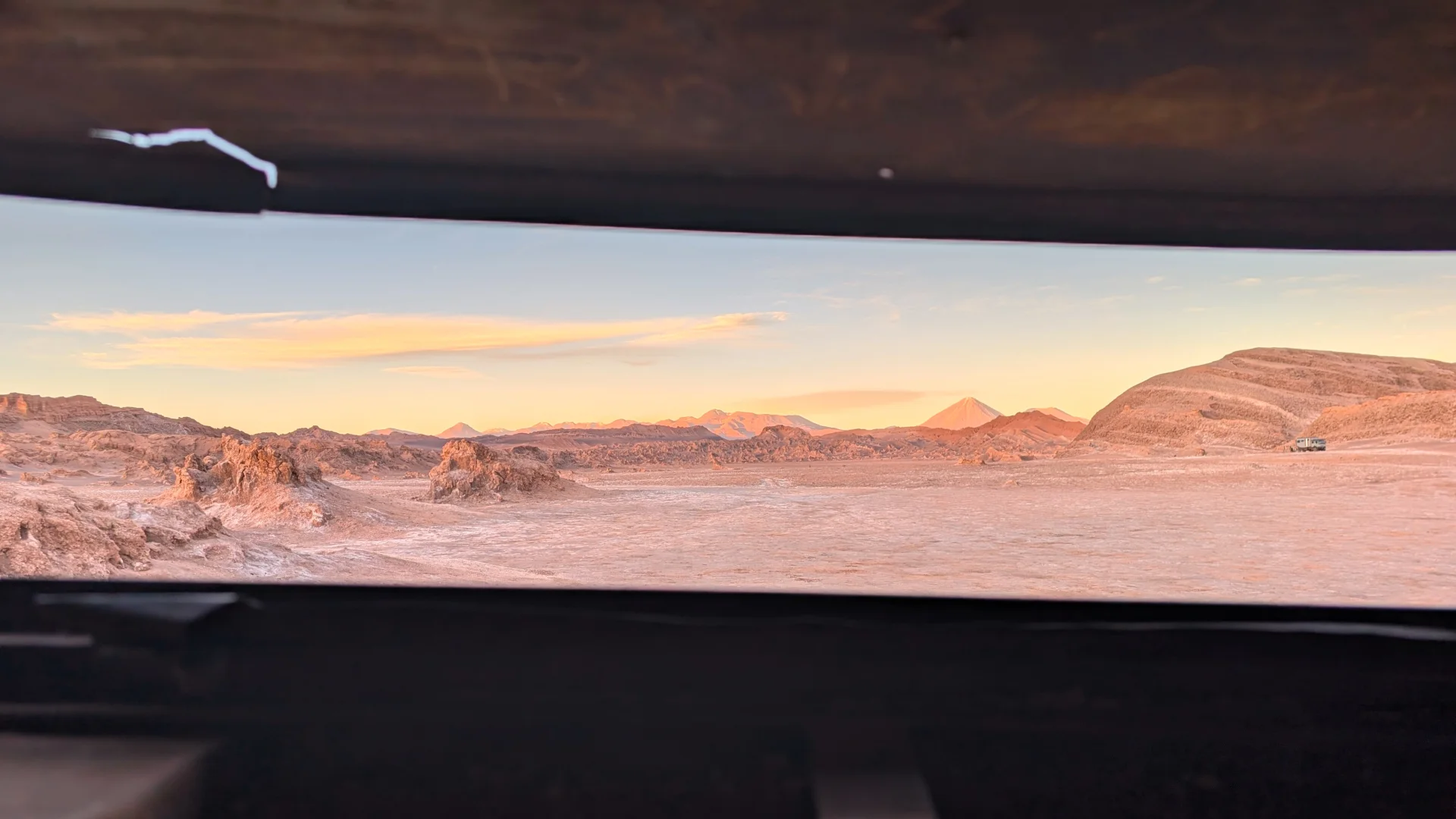 Vista del desert d'Atacama emmarcada a través d'una obertura horitzontal i fosca des de l'interior de l'autobús abandonat. A través de la fenedura es veu l'ampli paisatge de formacions rocoses i sorra de tons rosats, amb la silueta cònica del volcà Licancabur a l'horitzó sota un cel suau de posta de sol.