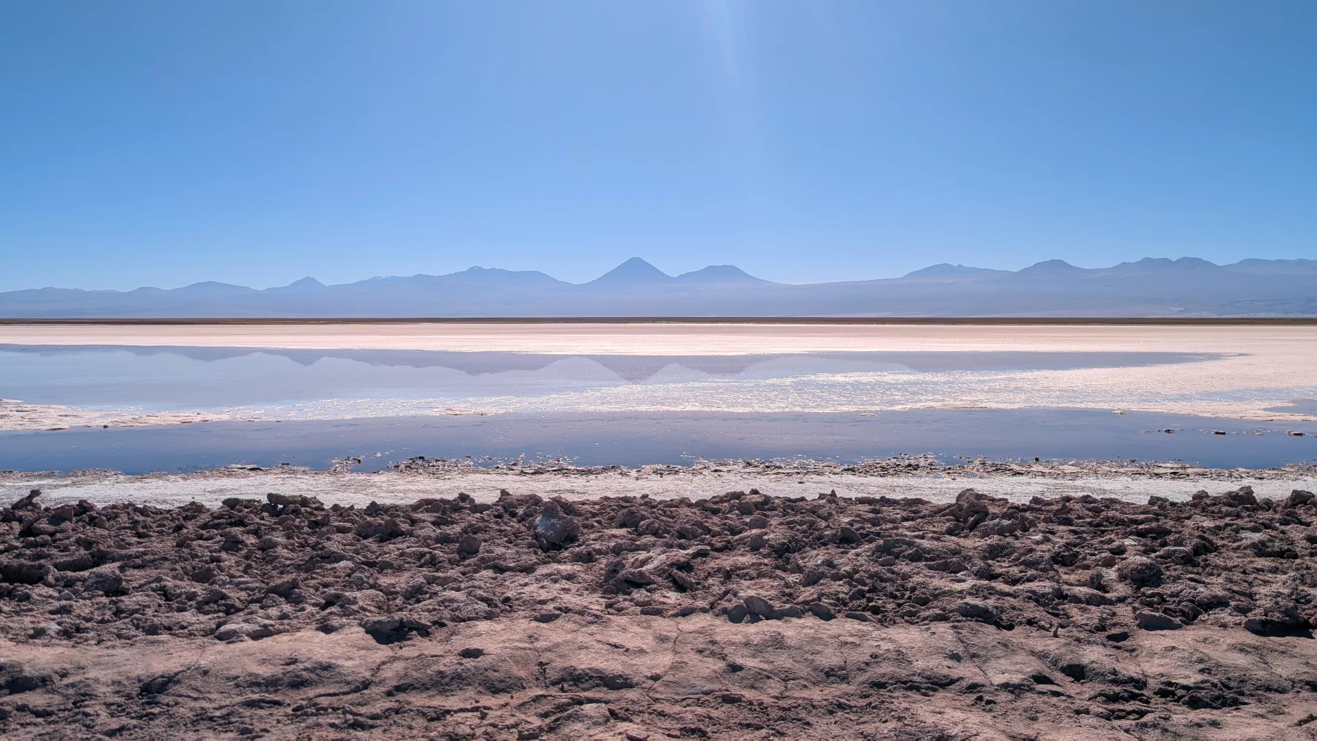 Vista de la Laguna Tebinquiche al desert d'Atacama, on una gran crosta de sal blanca i zones d'aigua poc profunda reflecteixen la serralada dels Andes a l'horitzó, sota un cel blau clar i radiant. Al primer pla es mostra el terreny àrid, terrós i rugós del salar.