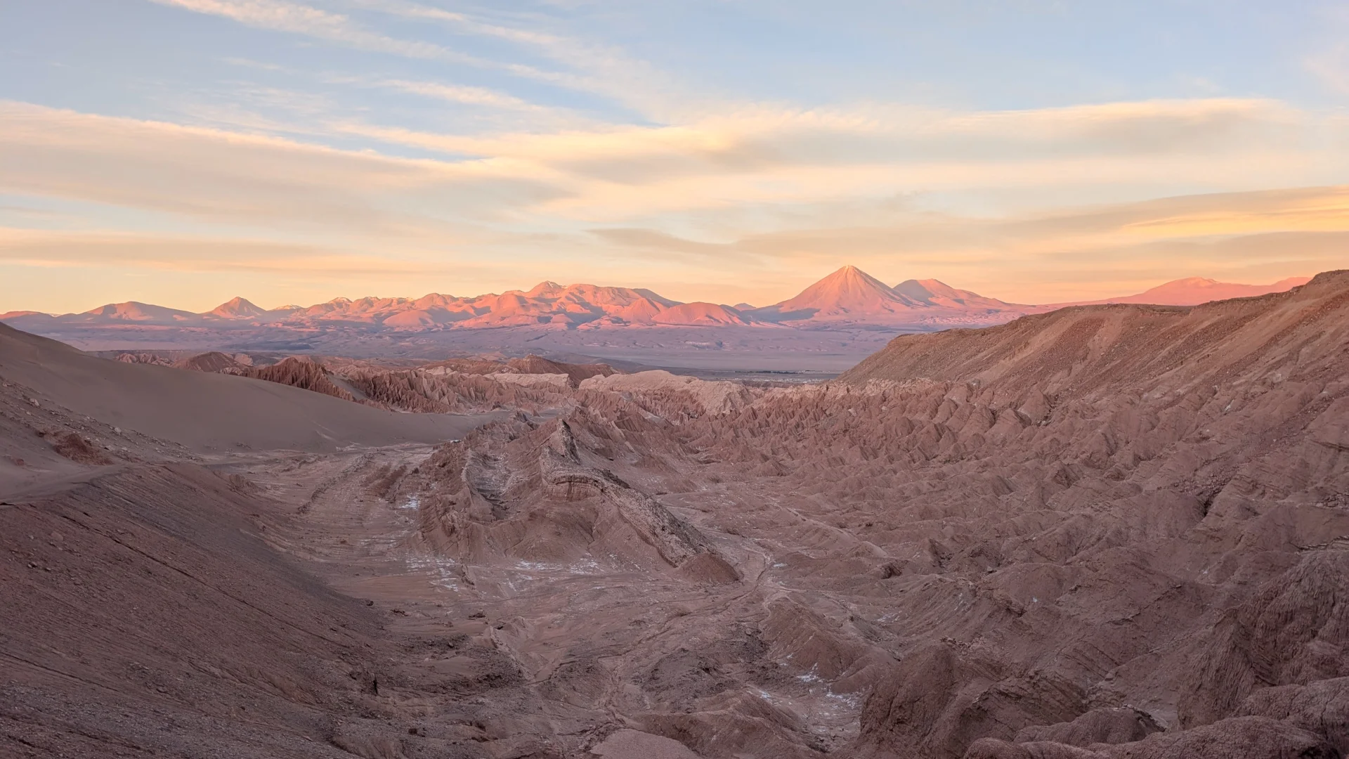 Vista panoràmica del Valle de Marte al desert d'Atacama durant la posta de sol, que mostra una vall escarpada amb formacions rocoses de tons marrons i una gran duna de sorra a la banda esquerra. Al fons, la serralada dels Andes i la silueta cònica del volcà Licancabur s'il·luminen amb suaus tons rosats sota un cel amb núvols prims de colors daurats.