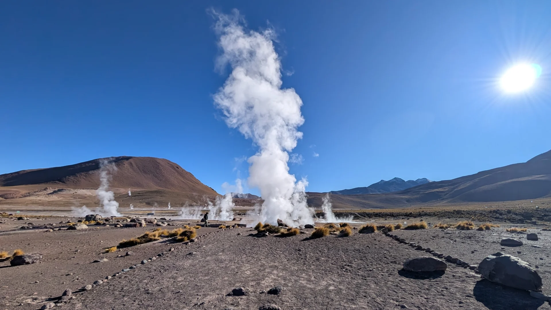Camp geotèrmic dels Guèisers del Tatio amb una gran i alta columna de vapor blanc que s'eleva verticalment des de terra. El terreny és àrid, de color marró i pedregós, amb camins delimitats per seguretat on es veu una silueta humana al fons. Al fons s'aixequen muntanyes pelades sota un cel blau clar amb el sol brillant de cara a la part superior dreta.