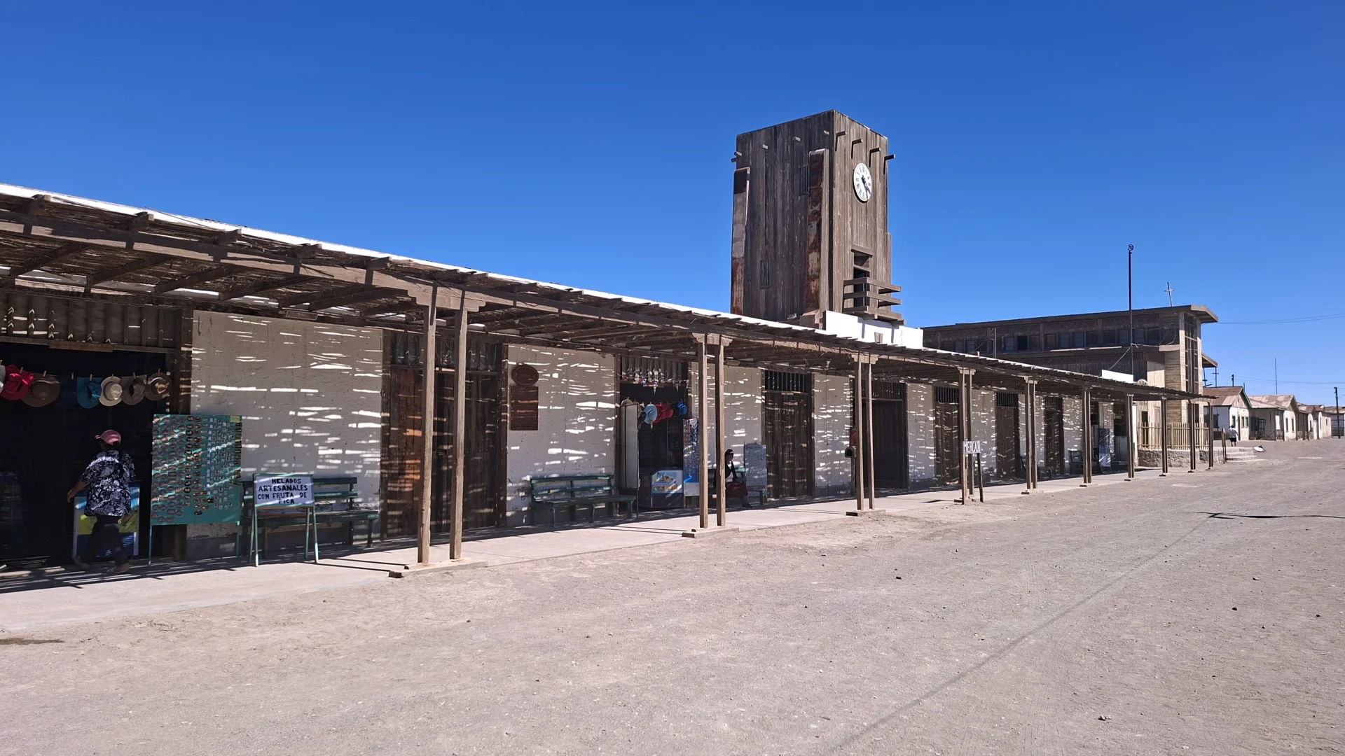 Vista de l'antiga oficina salnitrera de Humberstone, amb una llarga estructura blanca de planta baixa protegida per un gran porxo de fusta que projecta ombres allargades a la façana. Al fons destaca una característica torre de rellotge de forma quadrada construïda totalment de fusta fosca, tot envoltat per un ampli terra de terra desèrtica sota un cel blau clar i intens.