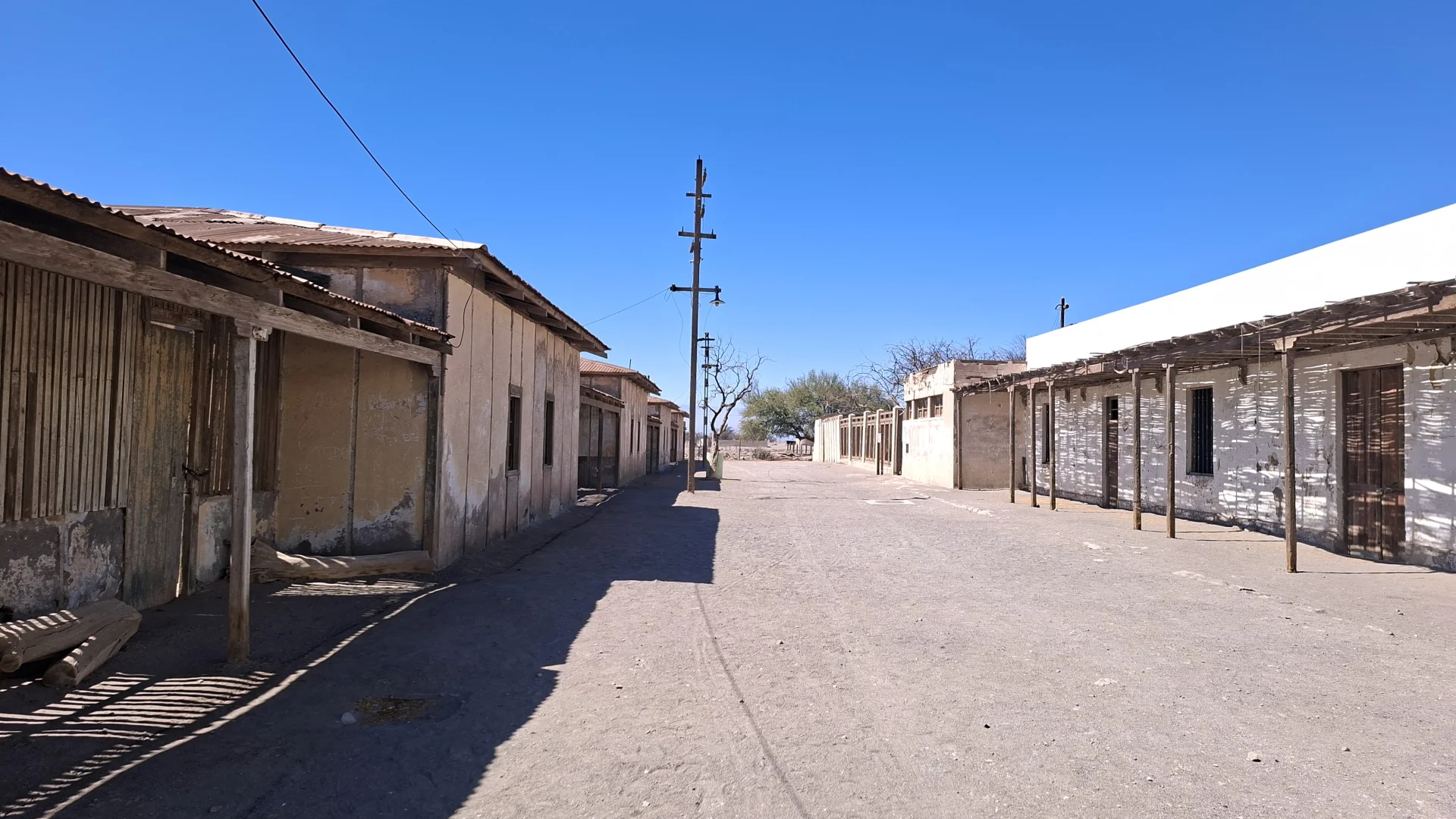 Carrer desert i polsegós a l'antiga colònia salnitrera de Humberstone, flanquejat a banda i banda per cases històriques d'una sola planta amb estructures i porxos de fusta que projecten ombres denses. Al mig del recorregut destaca un antic pal de fusta de línia elèctrica, tot sota la llum intensa del sol i un cel blau brillant sense núvols.