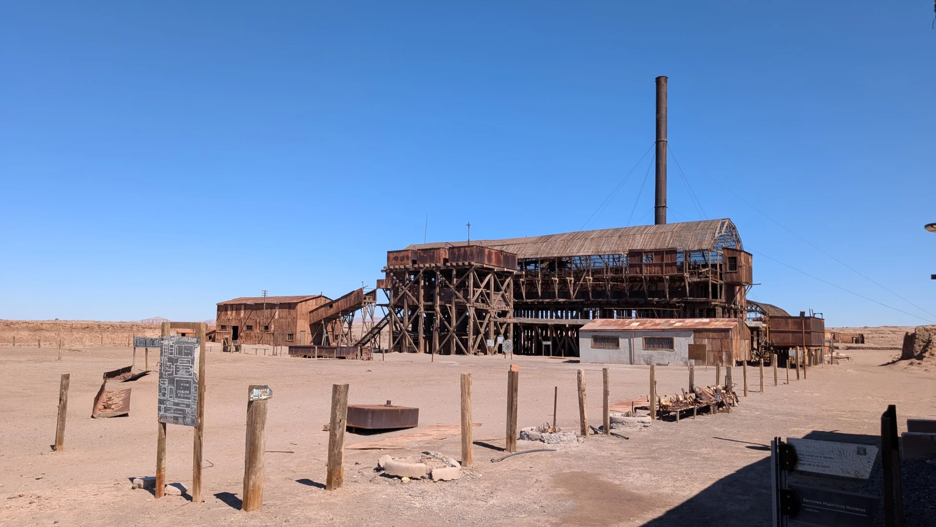 Vista de la gran planta de lixiviació de l'Oficina Santa Laura, una imponent estructura industrial abandonada de fusta fosca i ferro oxidat de diversos pisos, amb una llarga xemeneia que s'eleva cap al cel. El complex se situa en una esplanada de terra desèrtica amb pals de fusta, restes de maquinària i un panell informatiu en primer pla, sota un cel blau clar i intens.