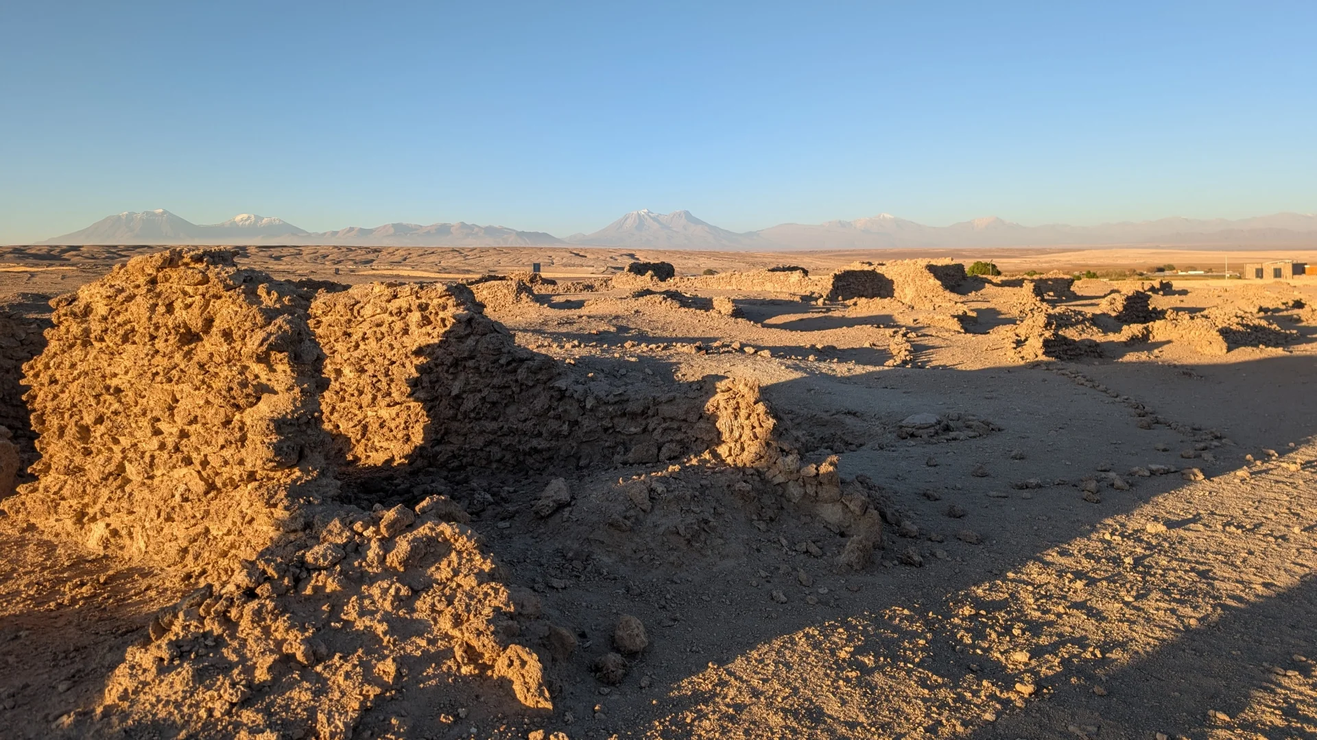 Vista de les ruïnes arqueològiques de pedra de la Pukará de Chiu Chiu envoltades d'un terreny desèrtic sota la llum càlida del sol, que projecta ombres allargades. Al fons, destaca una línia de muntanyes i volcans de l'altiplà amb els cims nevats a la llunyania sota un cel clar i net.