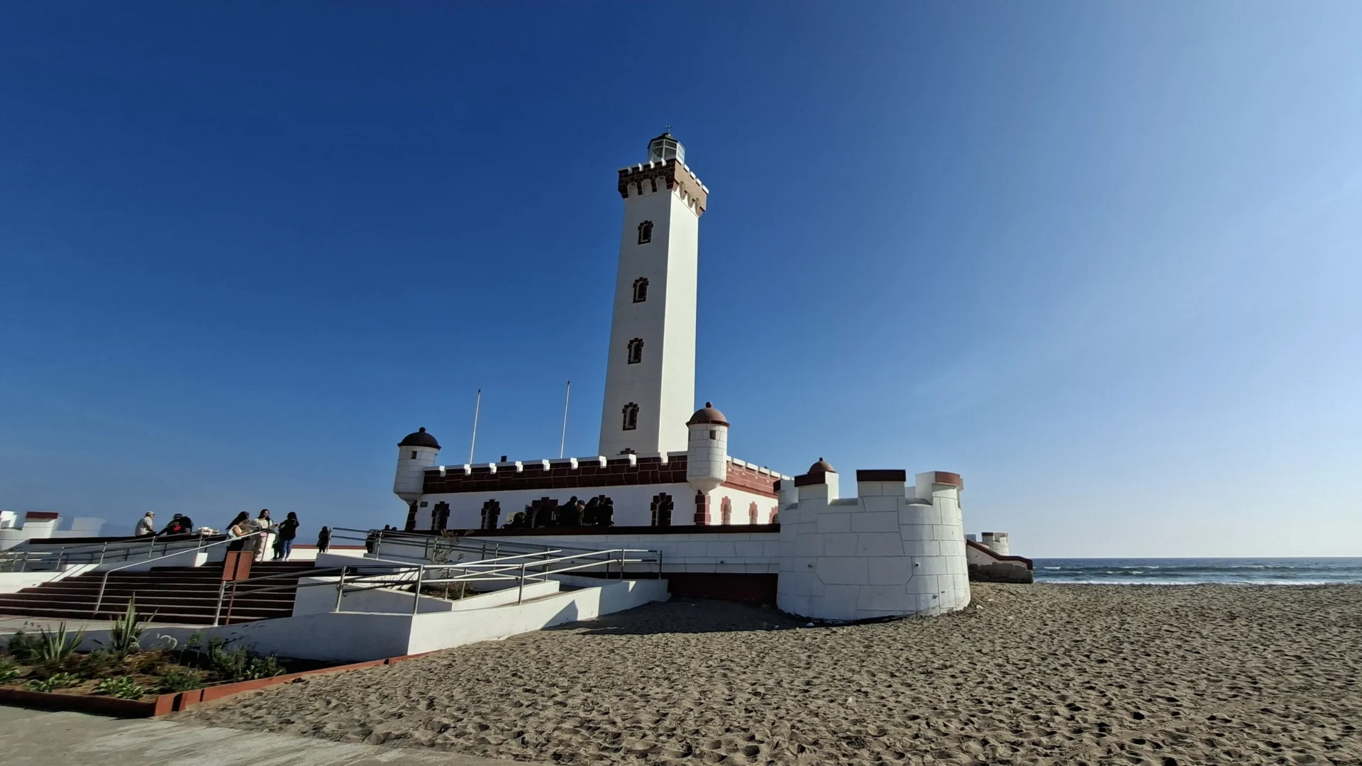 Una vista del Faro Monumental de La Serena, una estructura blanca i vermella amb elements de fortificació, situada a la platja sorrenca davant del mar blau sota un cel clar.