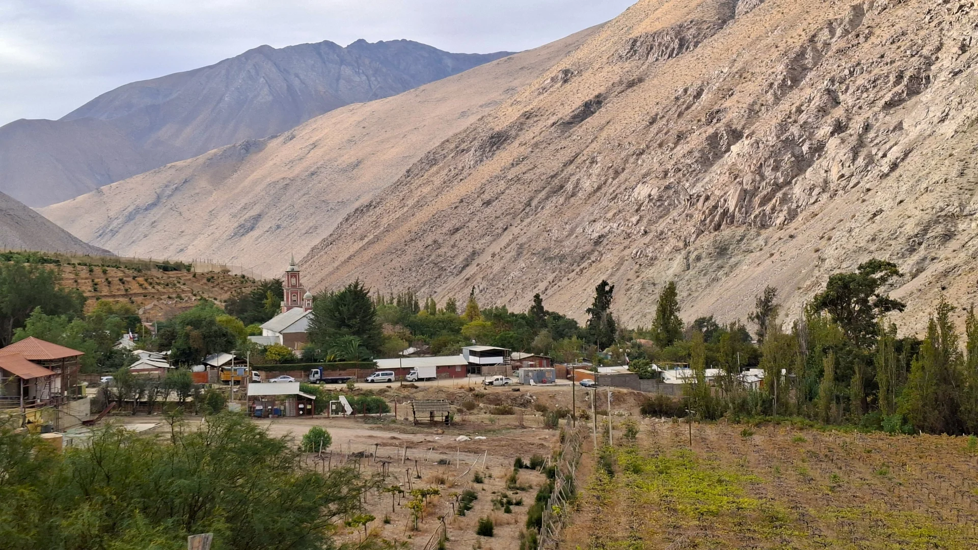 Una vista panoràmica de Pisco Elqui, un petit poble al Valle del Elqui, envoltat de vinyes verdes i arbres al peu de muntanyes àrides i escarpades sota un cel parcialment ennuvolat.