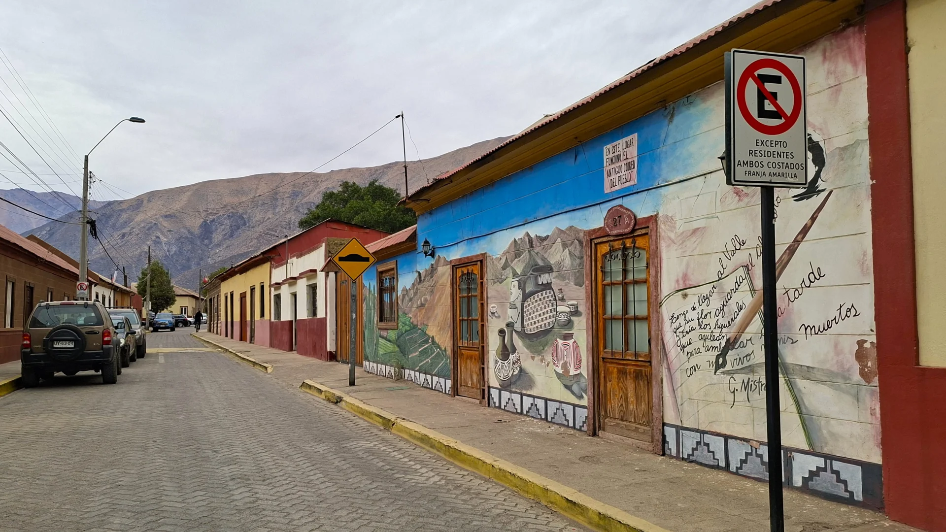 Una vista d'un carrer pintoresc a Diaguitas, Valle del Elqui, amb cases de colors i un gran mural detallat que cobreix la paret d'un edifici, sota un cel ennuvolat amb muntanyes al fons.
