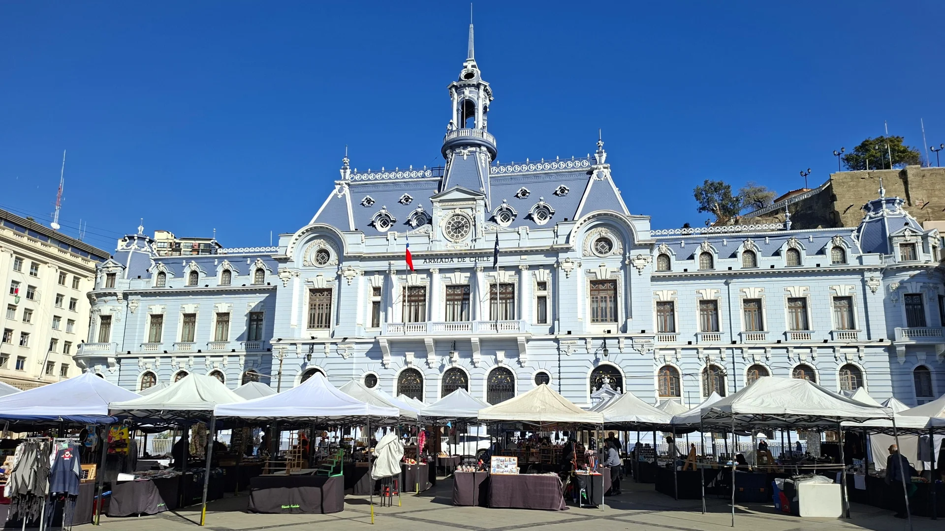 Vista frontal de l'edifici de la Comandància en Cap de l'Armada de Xile a Valparaíso, una imponent construcció d'estil clàssic de color blau cel amb una torre de rellotge central, amb un mercat de carpes blanques instal·lat a la plaça del davant sota un cel completament clar.