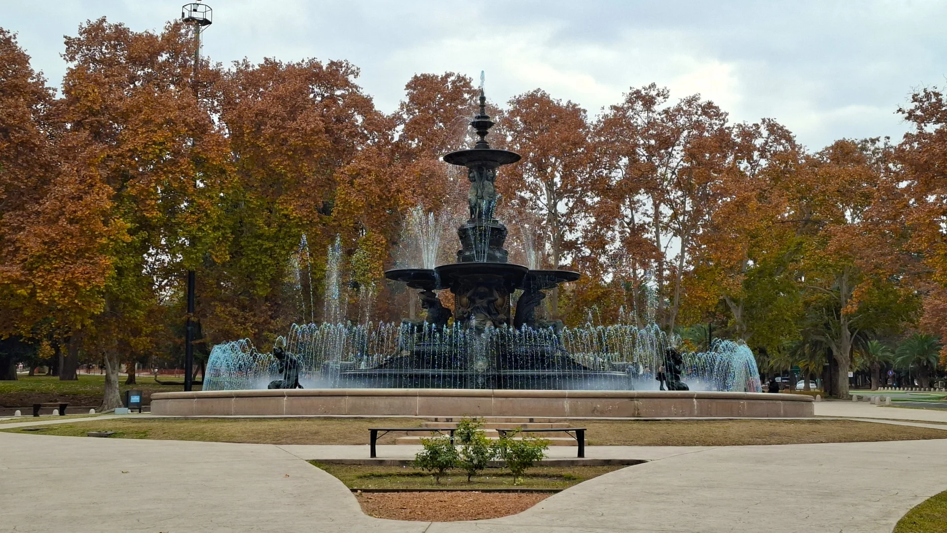 Gran font ornamental amb brolladors d'aigua en funcionament al Parc General San Martín de Mendoza, envoltada d'arbres frondosos amb fulles de tons tardorencs ocres i marrons sota un cel gris.