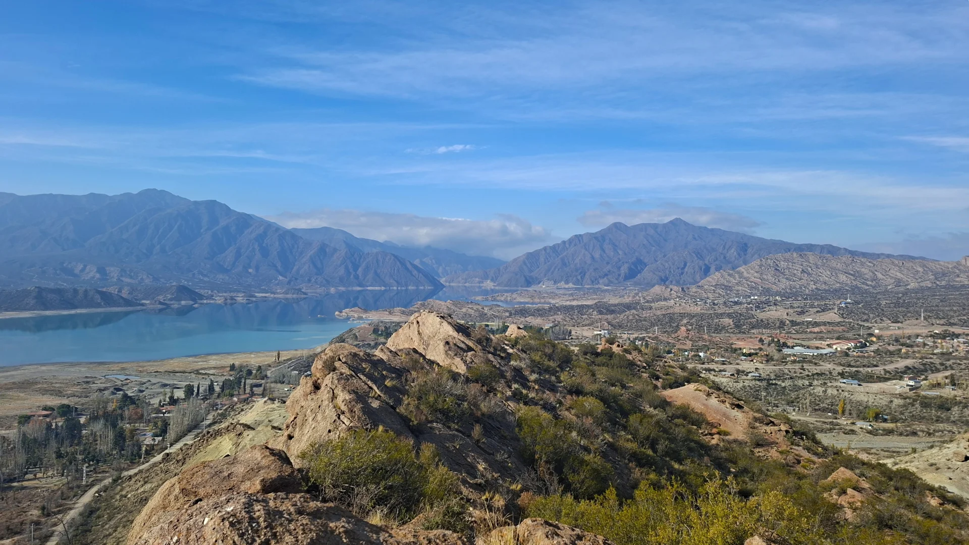 Vista panoràmica des del Cerro Cocodrilo a Potrerillos, on s'aprecia un gran embassament d'aigua blava reflectint els núvols i una vall àrida envoltada de cadenes muntanyoses sota un cel clar.