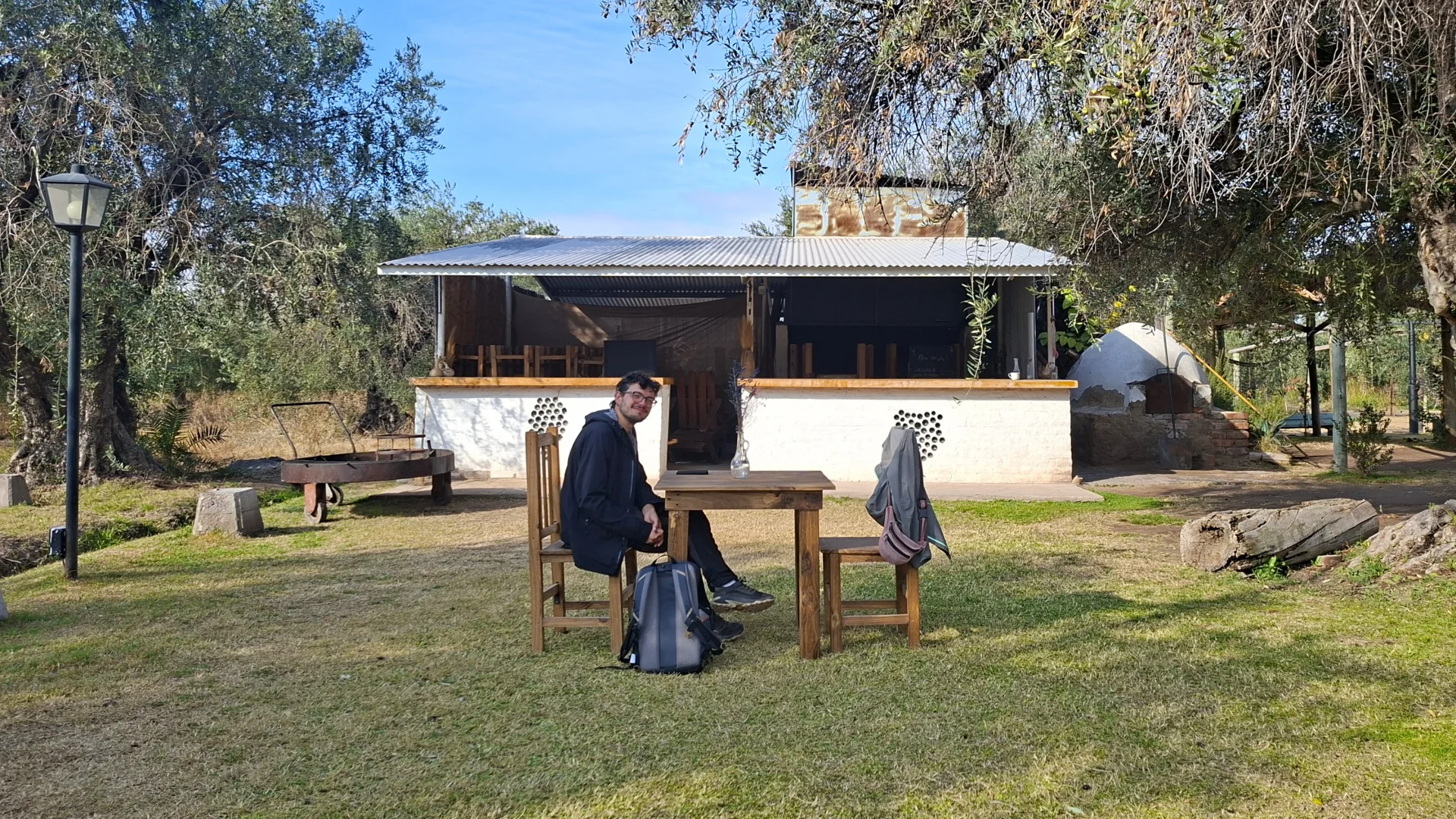 En Pau assegut en una cadira de fusta davant d'una taula en un jardí amb oliveres a la Bodega Sillon, amb una motxilla a terra i una edificació rústica blanca amb barra de fons.