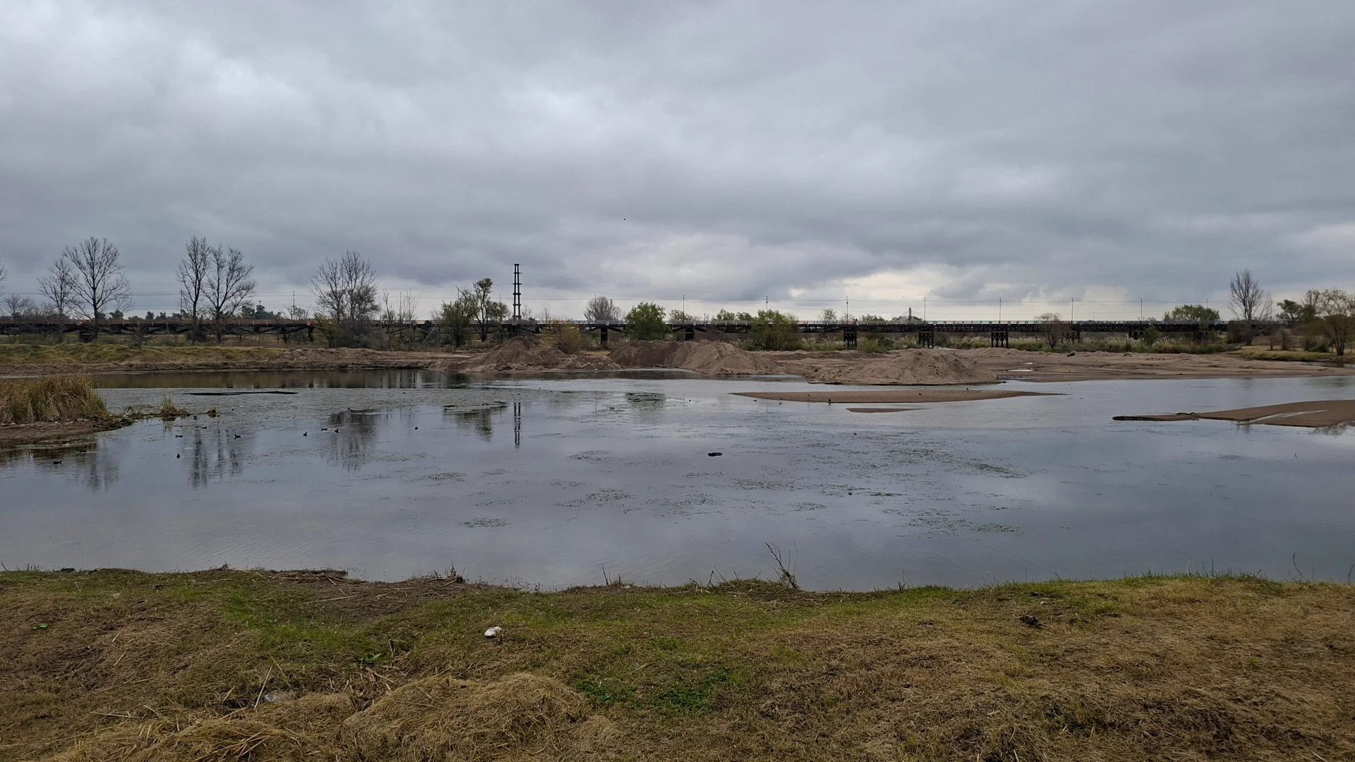 Vista del parc de la Costanera a Villa Mercedes sota un cel completament ennuvolat i gris, que mostra un riu ample i tranquil amb acumulacions de sorra i un llarg pont al fons.