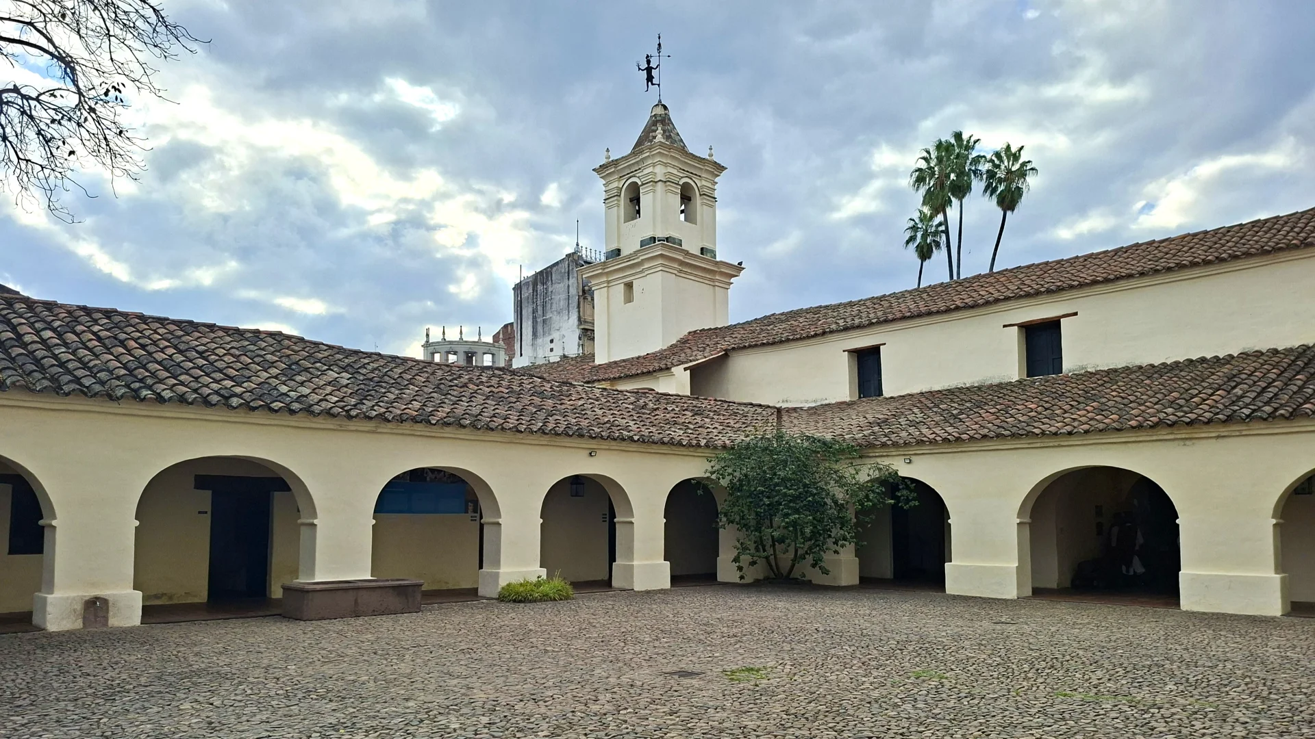 Vista del pati interior empedrat del Cabildo històric de Salta, envoltat per porxos amb arcs de mig punt emblanquinats sota una teulada tradicional de teula àrab, amb un petit arbre al centre i la torre blanca del campanar alçant-se al fons sota un cel amb núvols i palmeres.
