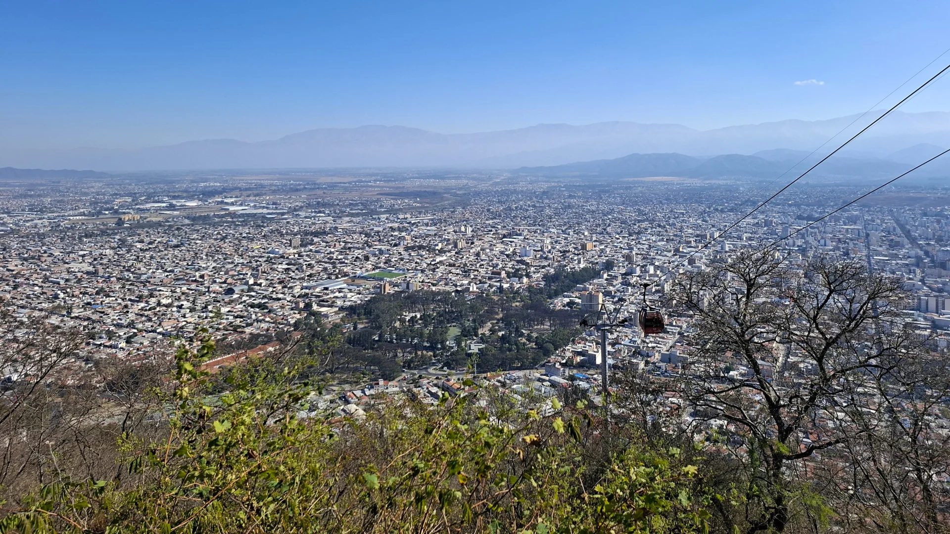 Vista panoràmica de la ciutat de Salta des del mirador del Cerro San Bernardo sota un cel blau clar, mostrant l'extens traçat urbà que s'estén per tota la vall fins a les muntanyes de l'horitzó, amb la vegetació del vessant i una cabina vermella del telefèric suspesa dels cables a la dreta.