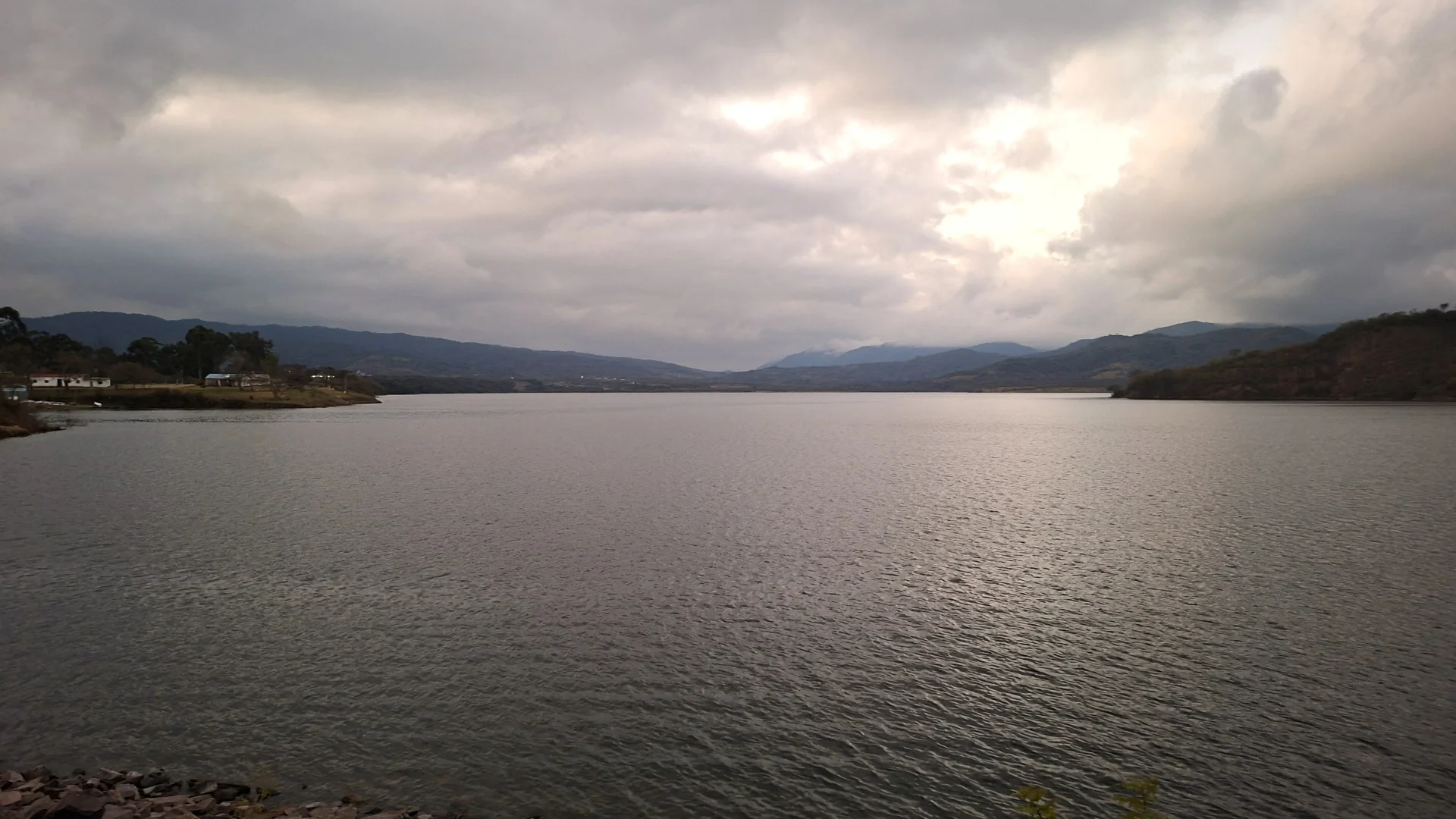 Vista panoràmica de la represa de Campo Alegre al capvespre, amb les aigües tranquil·les de l'embassament estenent-se sota un cel cobert de núvols grisos amb suaus reflexos de llum, flanquejades per turons i muntanyes a l'horitzó.