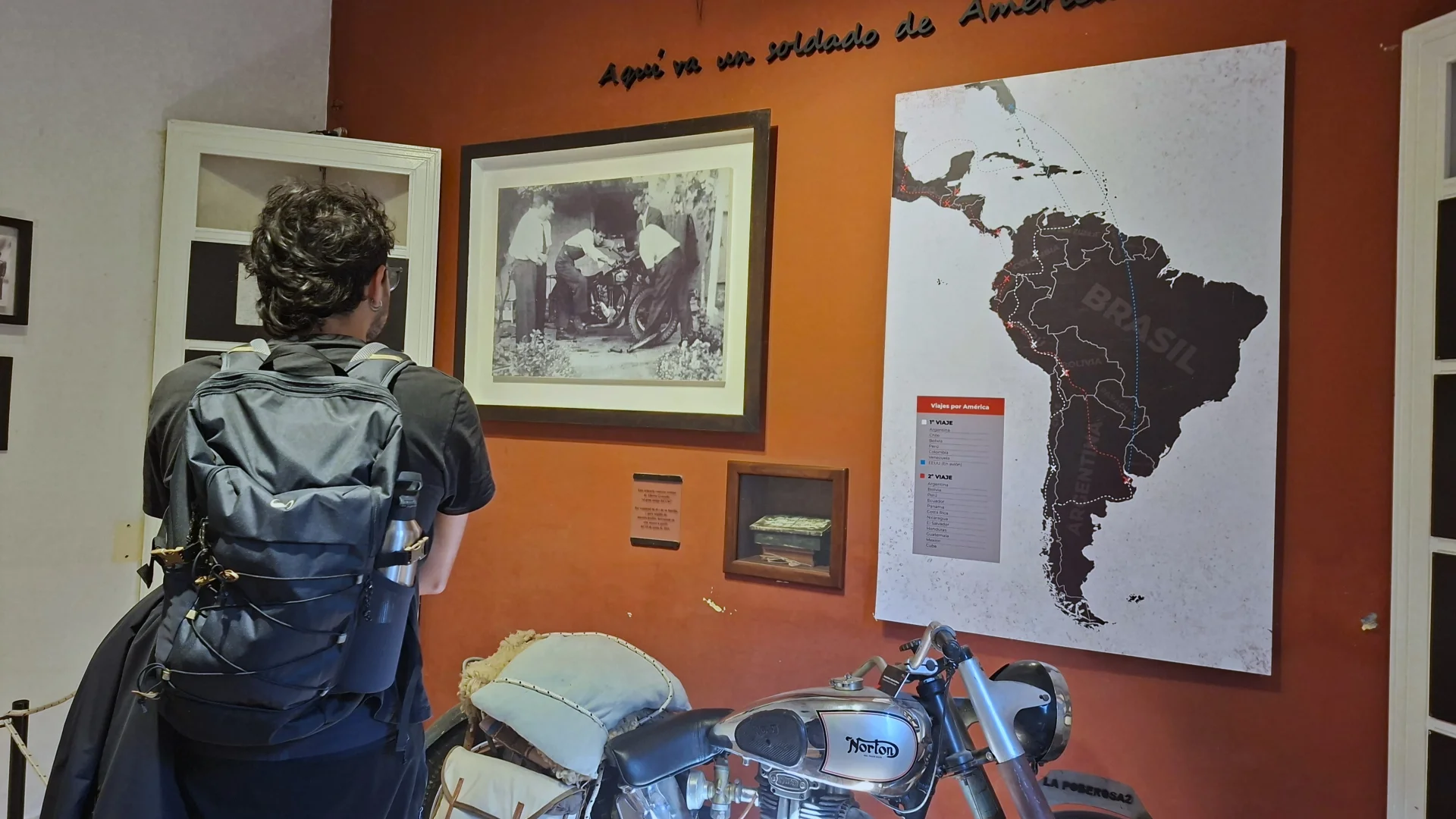Un visitant d'esquena amb motxilla negra observa una exposició a l'interior del Museu Casa d'Ernesto Che Guevara a Alta Gracia, que compta amb una paret vermella decorada amb un mapa de l'Amèrica del Sud i fotografies històriques, i amb la rèplica de la motocicleta Norton 'La Poderosa II' en primer pla.