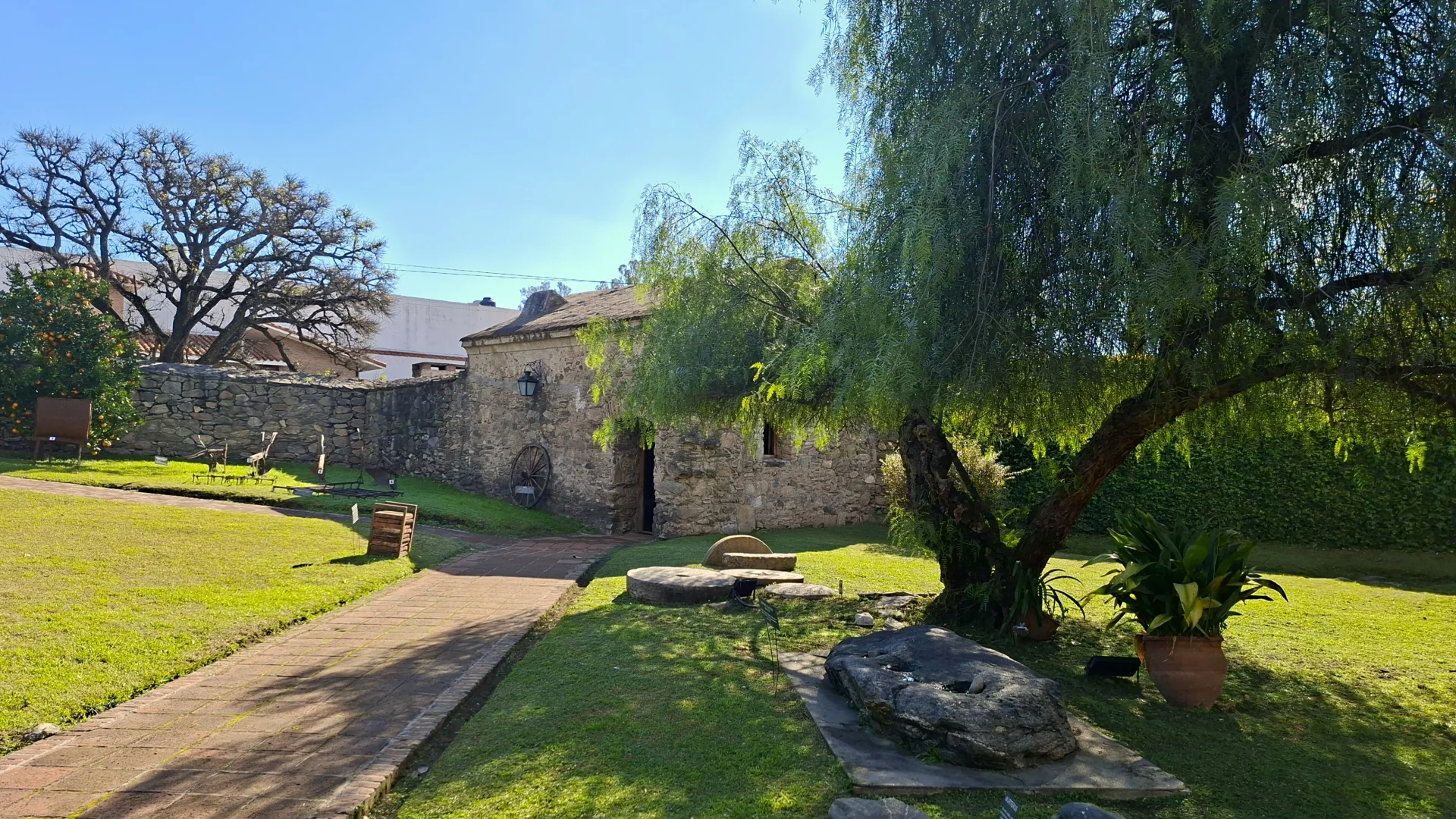 Un pati interior enjardinat de l'Estància Jesuítica d'Alta Gracia, que mostra una antiga edificació de pedra, un gran arbre que fa ombra sobre la gespa verda, un camí de maons i una antiga roda de carro de fusta recolzada a la paret.