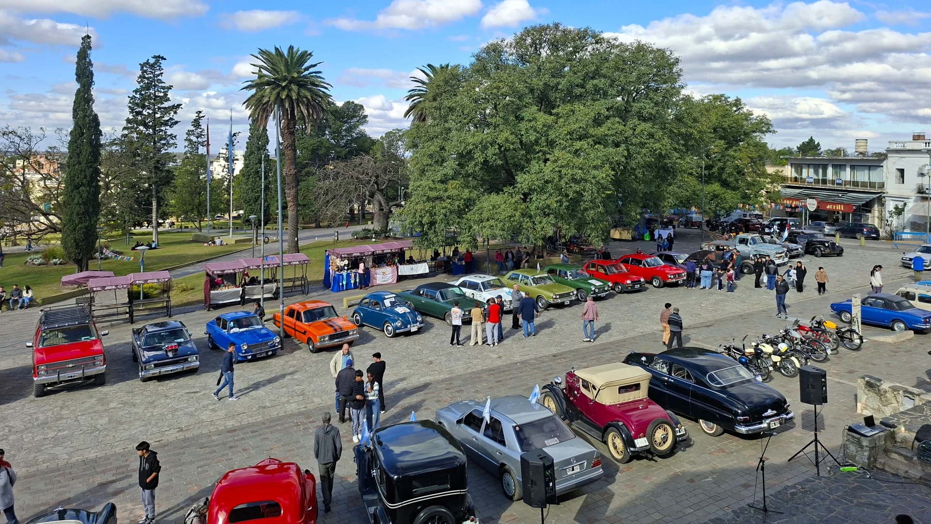 Vista elevada d'una exposició de cotxes i motocicletes clàssiques de diversos colors en una plaça pública d'Alta Gracia, envoltada de palmeres, grans arbres i persones passejant sota un cel blau amb núvols blancs.