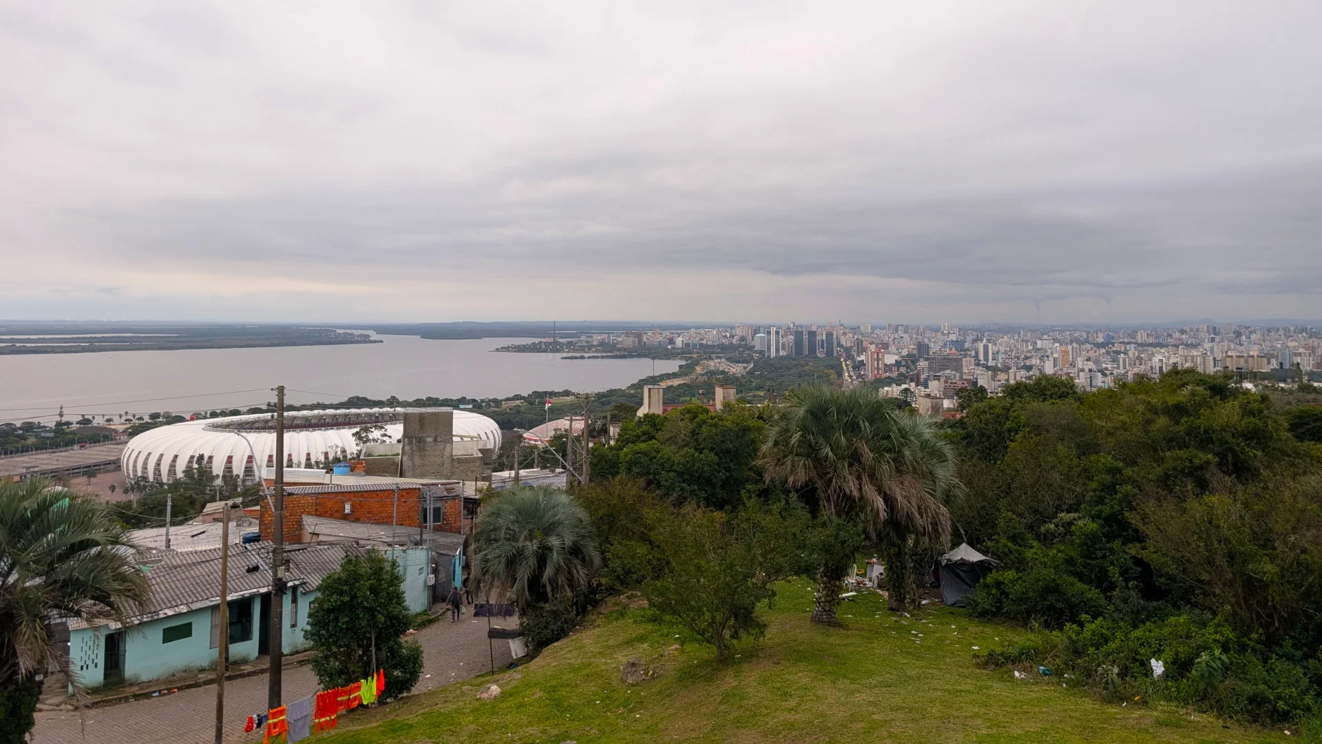 Vista panoràmica i elevada de la ciutat de Porto Alegre sota un cel gris i ennuvolat, on es pot veure l'estructura blanca de l'estadi Beira-Rio a l'esquerra, el riu Guaíba i l'horitzó ple d'edificis urbans.