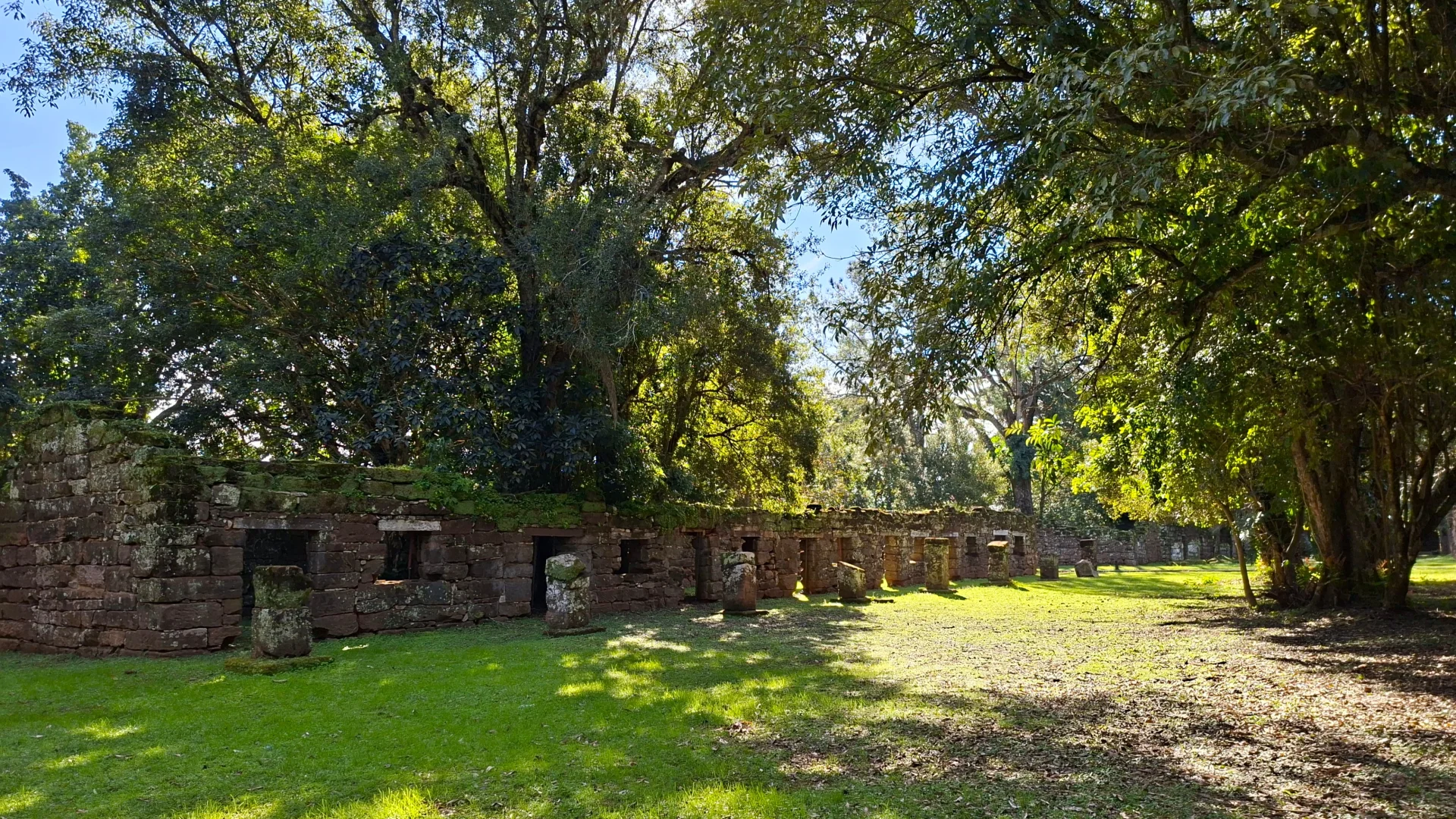 Una filera de parets estructurals de pedra antiga pertanyents a les ruïnes de San Ignacio Miní, envoltades d'una gran esplanada de gespa verda i sota l'ombra de grans arbres frondosos.
