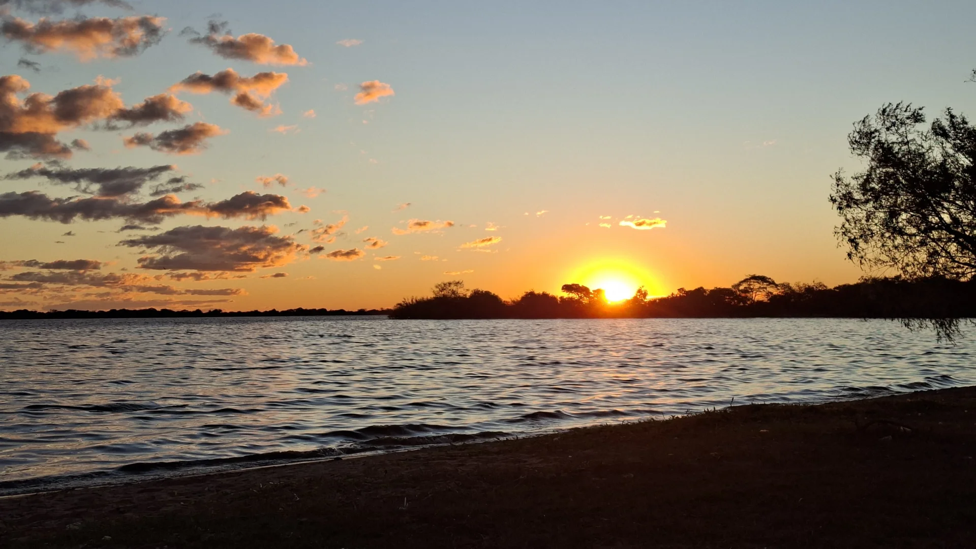 Posta de sol sobre el llac Guaíba a Porto Alegre, amb el sol ponent-se a l'horitzó entre siluetes d'arbres i núvols il·luminats amb tons taronges i daurats que es reflecteixen en l'aigua.