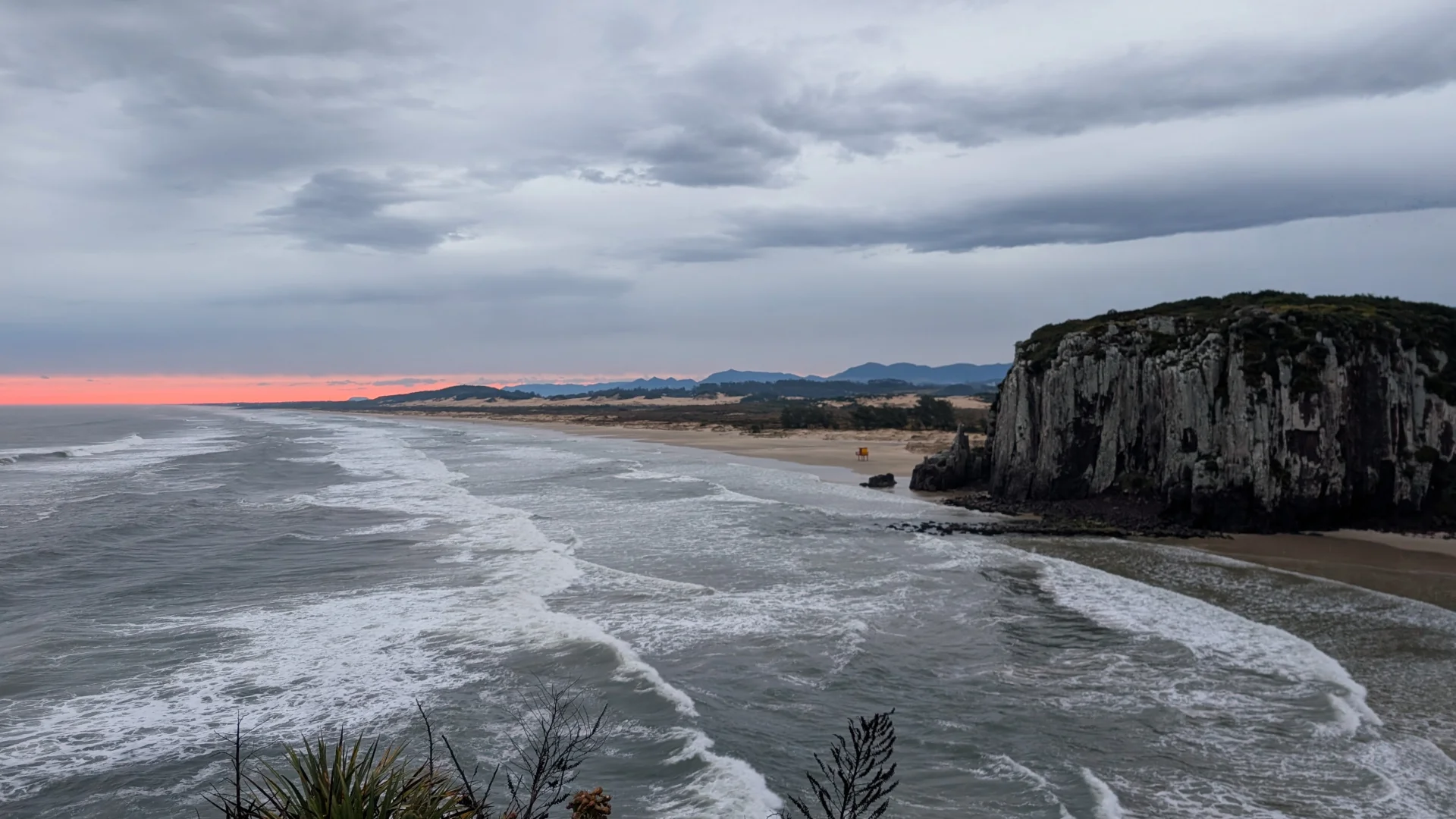 Vista panoràmica de la Praia da Guarita a Torres, amb ones del mar rompent a la sorra de la platja, un gran penya-segat rocós a la dreta i una franja taronja de la posta de sol a l'horitzó sota un cel ennuvolat.