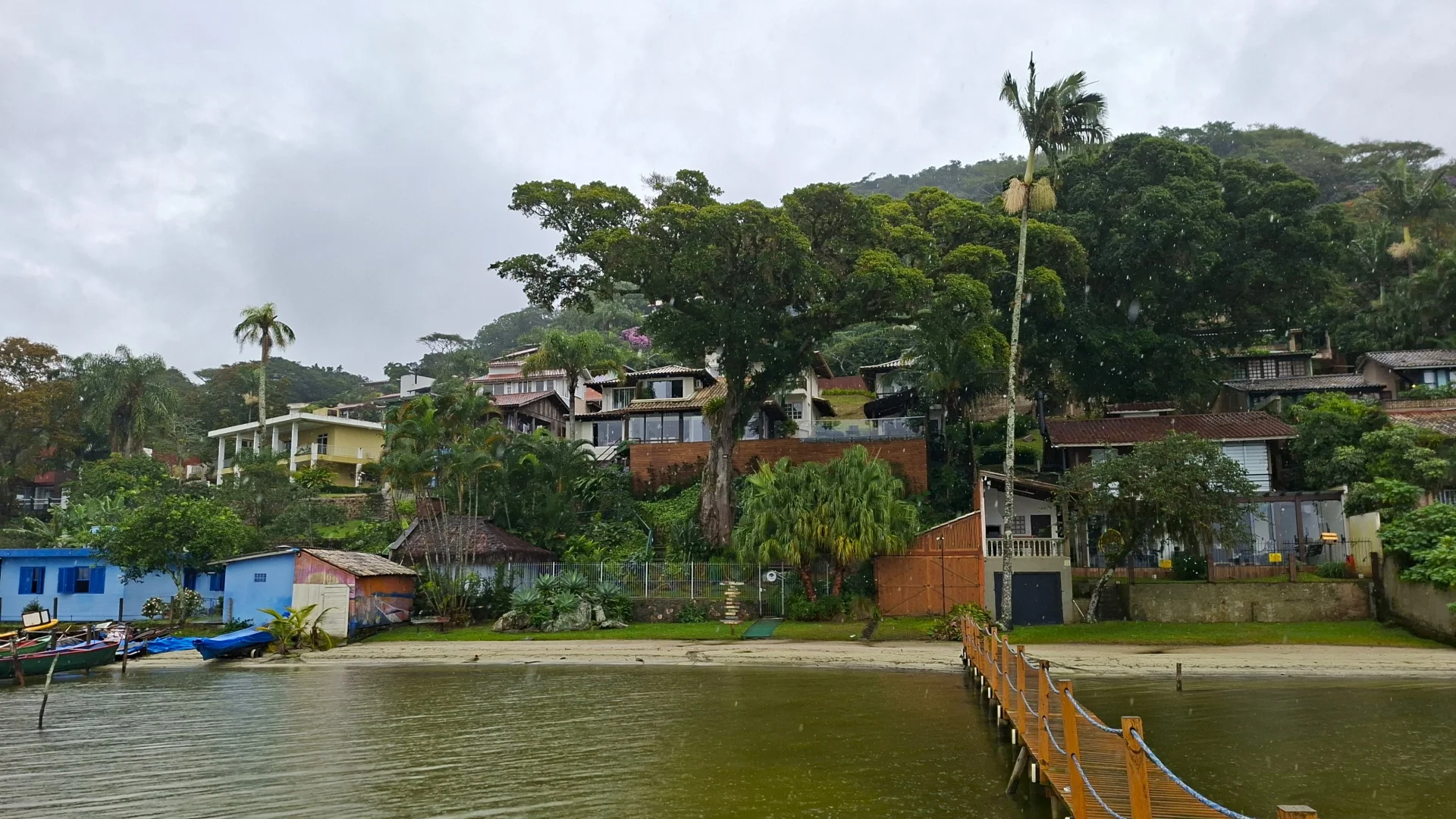 Una vista des de l'aigua que mostra un poble costaner de la Costa da Lagoa, a l'illa de Florianópolis, amb cases acolorides i edificis integrats en una muntanya densament coberta per bosc tropical i selva, amb un moll de fusta a la dreta que s'endinsa a l'aigua.