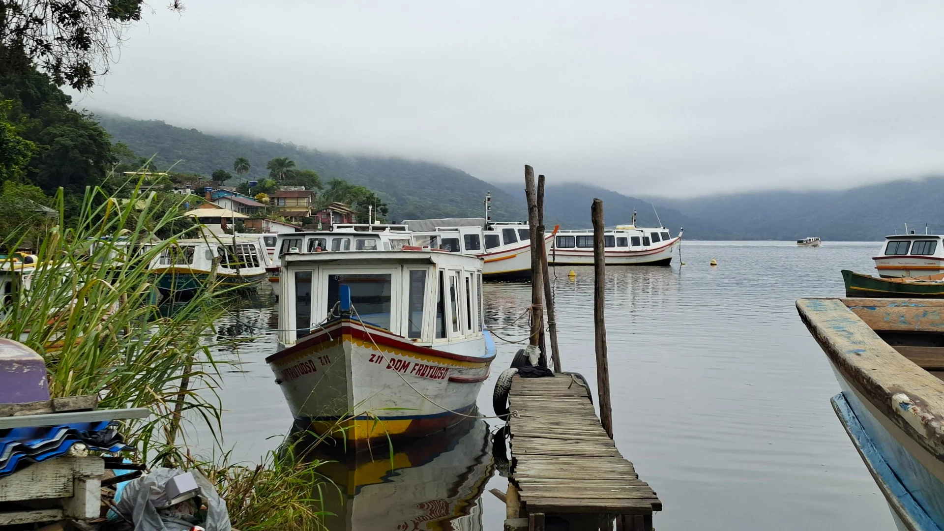 Una vista des de l'embarcador de fusta cap al moll de la Costa da Lagoa en un dia de pluja, que mostra moltes barques de pescadors de colors blau, groc i blanc, amarrades a la vora de l'aigua a prop del poble i les muntanyes cobertes per una espessa boira.