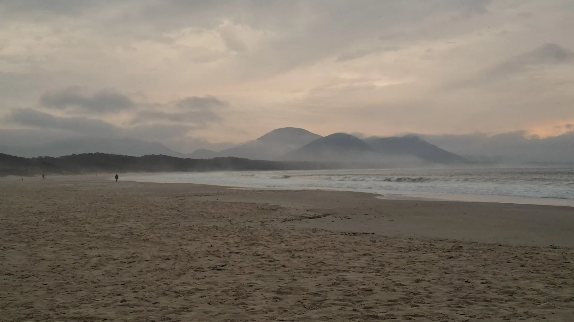Una vista panoràmica de la platja al capvespre sota un cel gris i ennuvolat, on es veuen les ones de l'oceà Atlàntic que es trenquen suaument a la sorra humida i deserta, amb la silueta d'una persona al fons caminant per la costa i muntanyes al fons enquadrades per la boira.