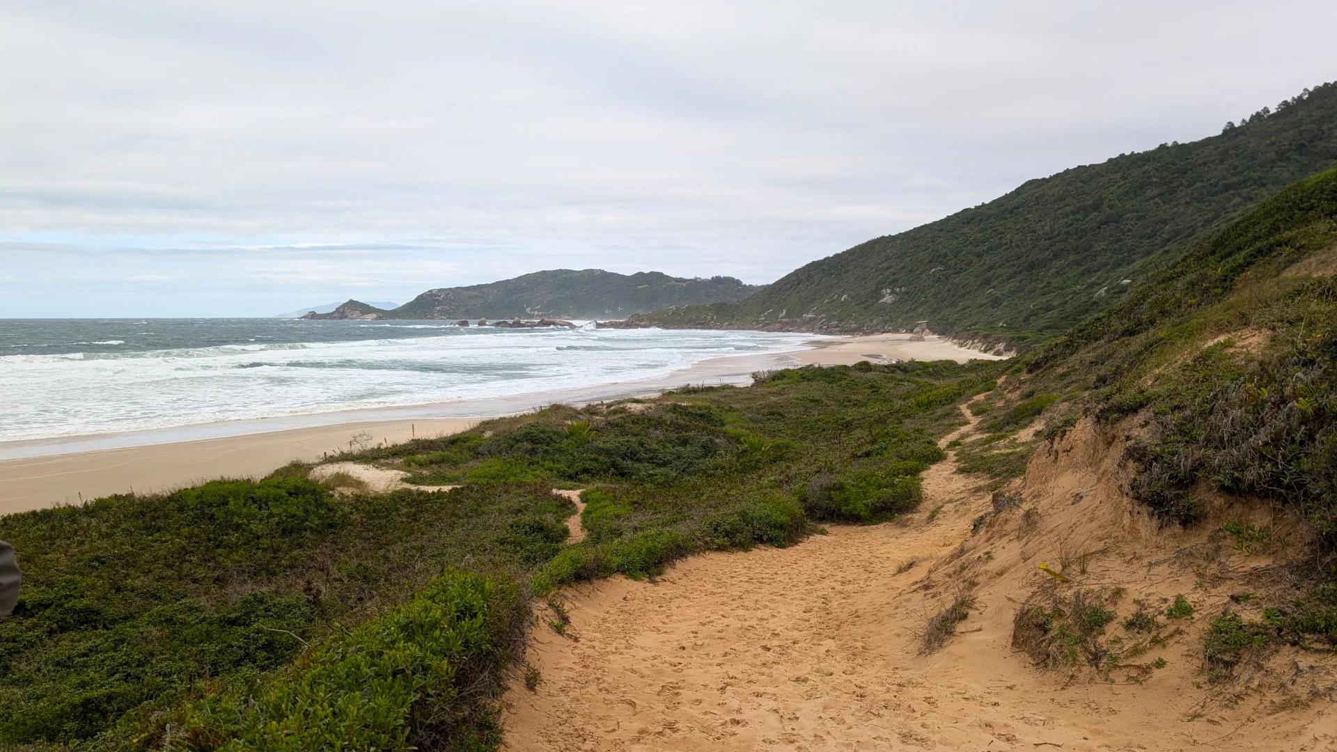 Una vista frontal d'un camí de sorra sinuós que porta avall cap a una gran platja sorrenca, amb les ones de l'oceà Atlàntic trencant a la vora. El camí està flanquejat per matolls baixos i, a la dreta, per un turó escarpat cobert de bosc tropical sota un cel ennuvolat i gris.