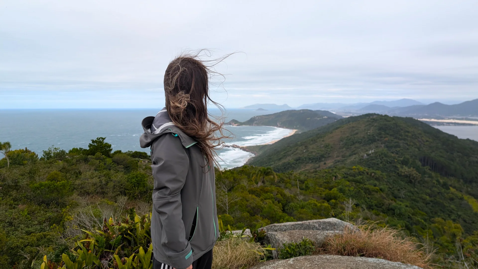 Una dona vista de costat i d'esquena des d'un punt de vista alt sobre una serra, amb els cabells ventats, portant una jaqueta impermeable de color gris i mirant cap a l'oceà. A sota d'ella s'estén una vasta cadena muntanyosa coberta de bosc tropical dens, una gran badia amb platges de sorra i turons al fons sota un cel ennuvolat.