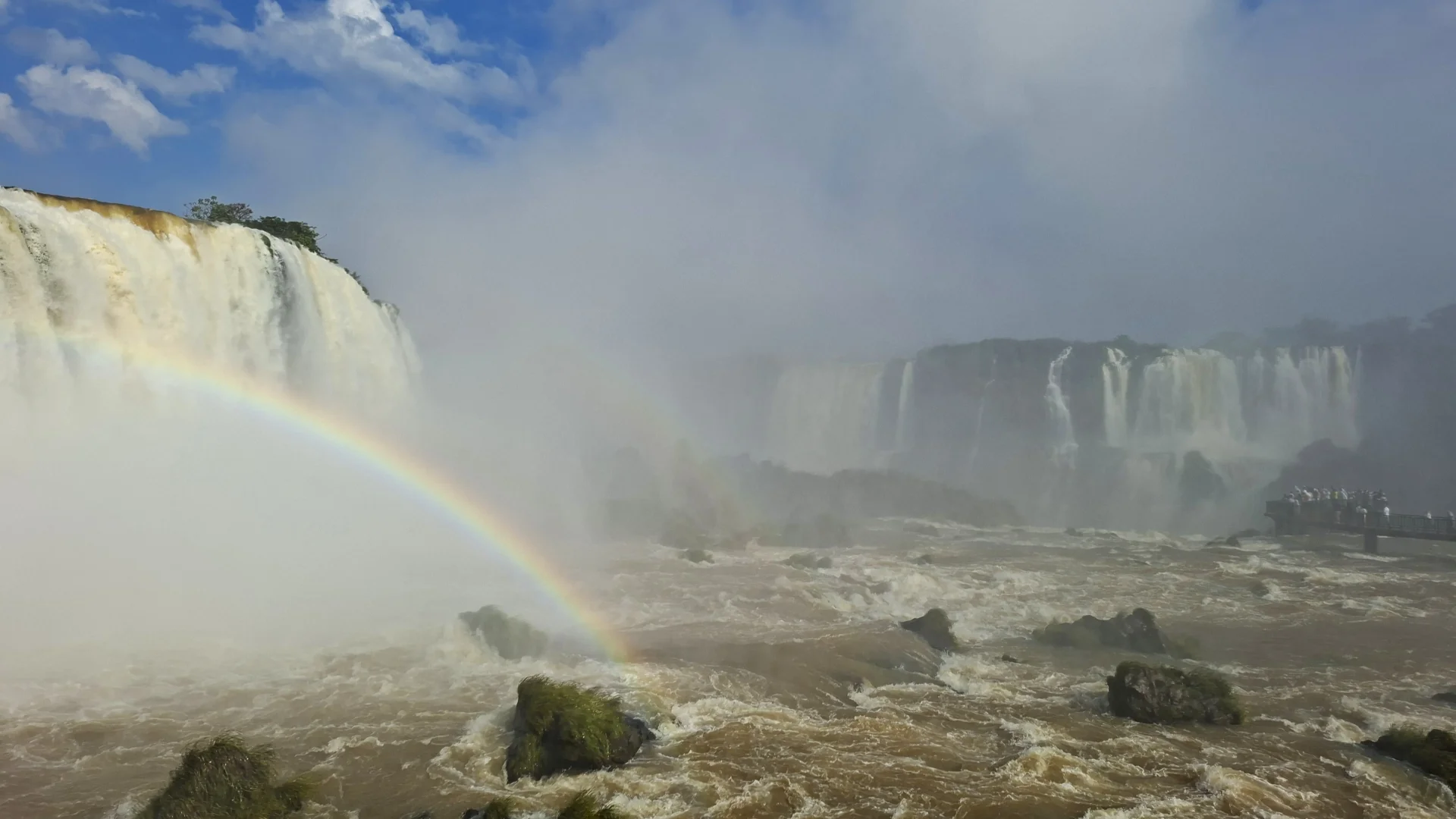 Vista de les Cascades de l'Iguaçú des del cantó brasiler, amb un arc de Sant Martí brillant que creua la boirina d'aigua en primer pla i grans salts d'aigua al fons sota un cel blau amb núvols.