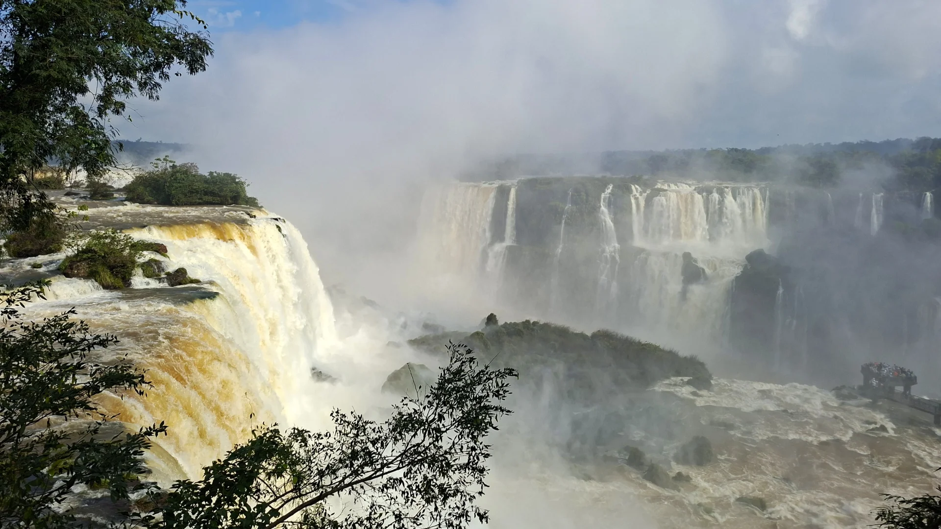 Imponent vista de la força de les Cascades de l'Iguaçú, amb grans columnes d'aigua caient que aixequen un dens núvol de boira blanca, amb vegetació en primer pla i una passarel·la amb visitants al fons.