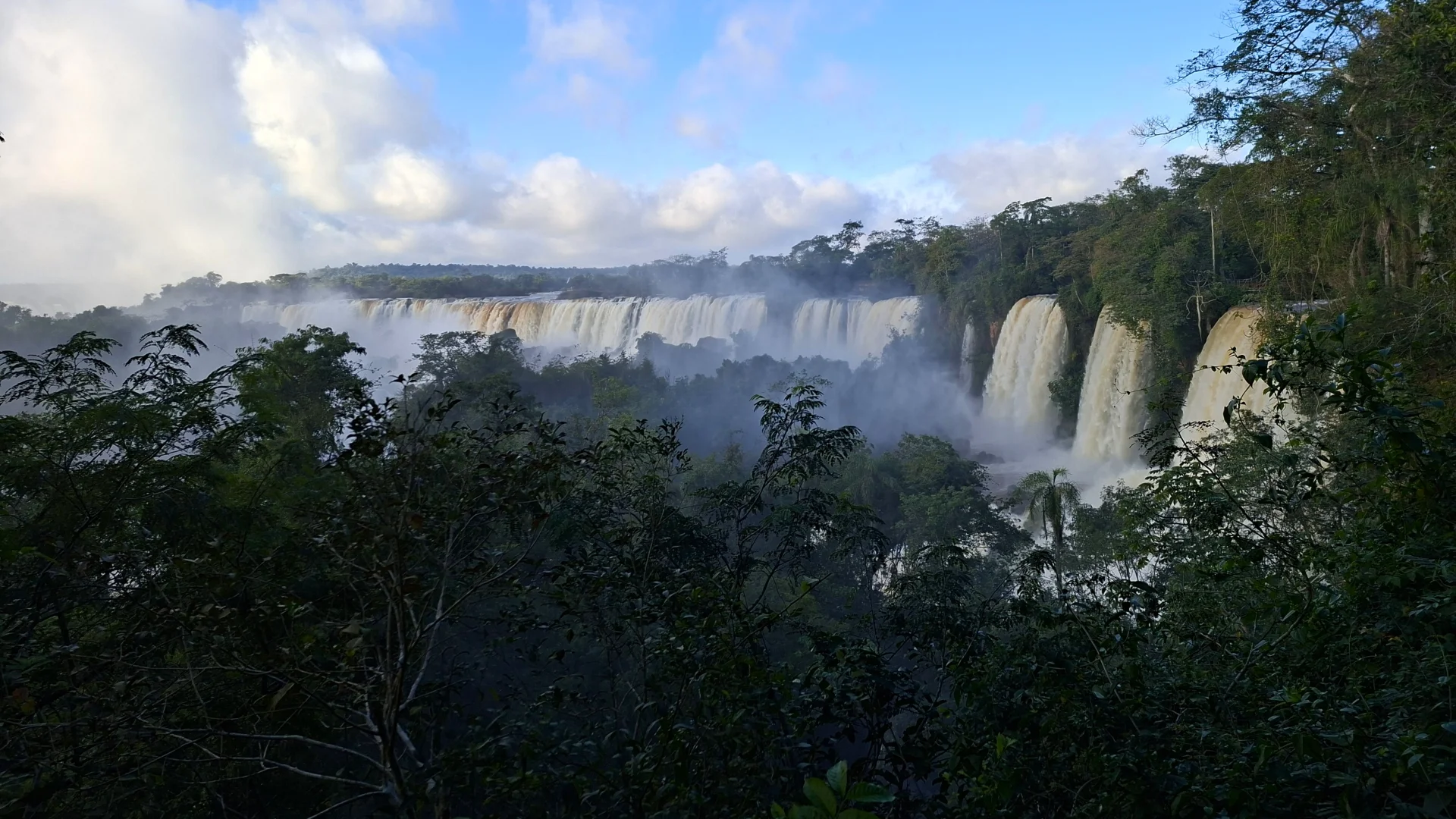 Vista panoràmica de les Cascades de l'Iguaçú des del cantó argentí, emmarcada per les branques i fulles de la selva tropical en primer pla, amb diversos salts d'aigua amples sota un cel blau amb núvols.