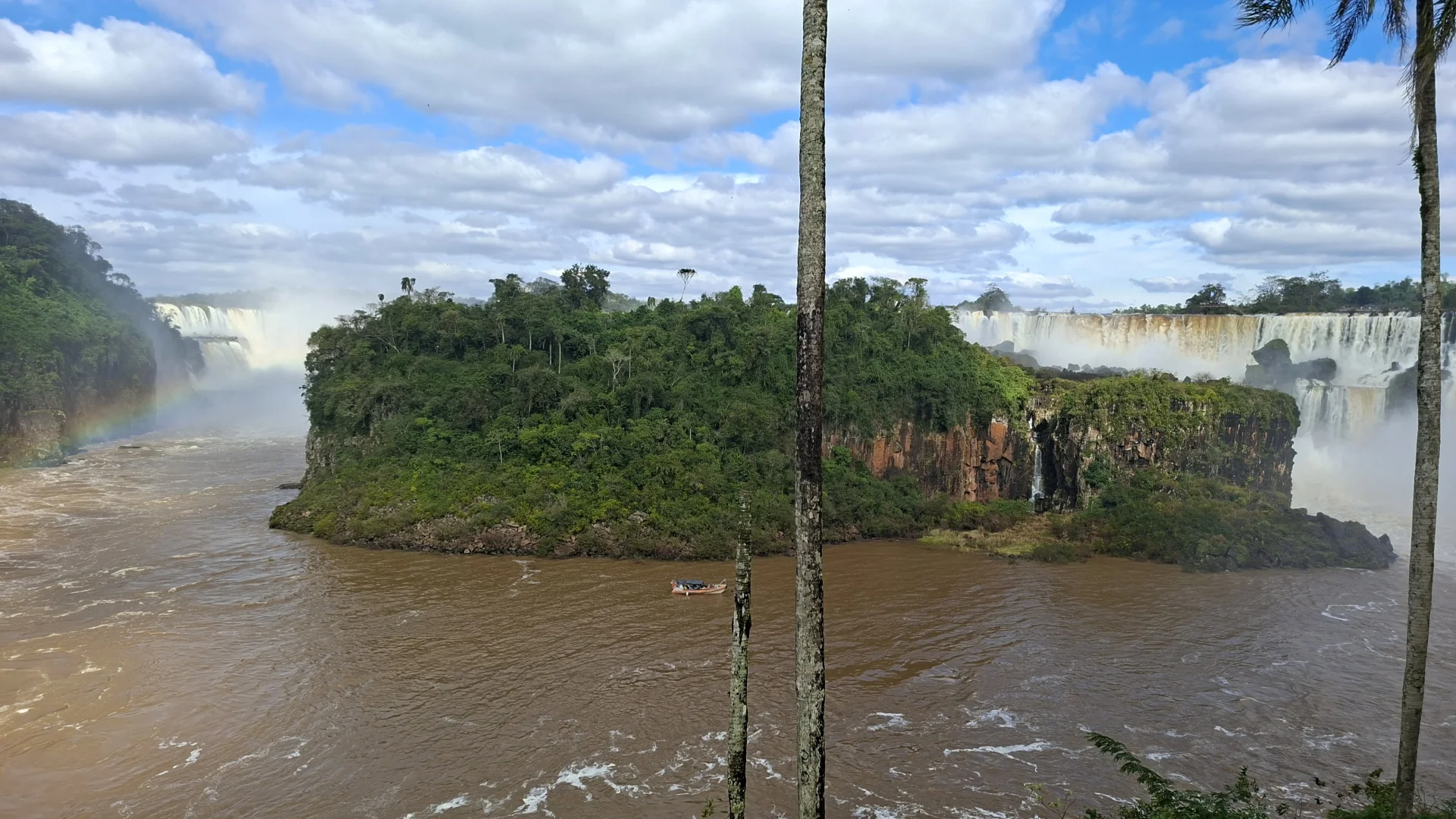 Una àmplia vista panoràmica de les imponents Cascades de l'Iguaçú, dividides per una gran illa central frondosa i plena de selva. A l'esquerra, un arc de Sant Martí intens es dibuixa sobre l'aigua caient; a la dreta, una llarga franja de salts d'aigua es precipita pels penya-segats. L'aigua marró i moguda del riu envolta l'illa, per on navega una petita embarcació, amb dos troncs prims de palmera emmarcant el costat dret sota un cel blau amb núvols.